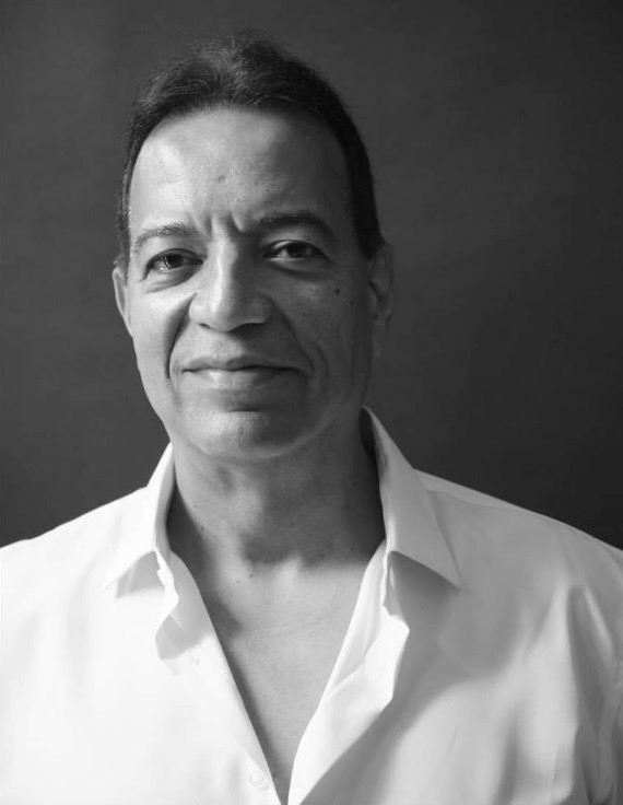Portrait of a man with short dark hair, wearing a white shirt, looking at the camera, against a dark background.