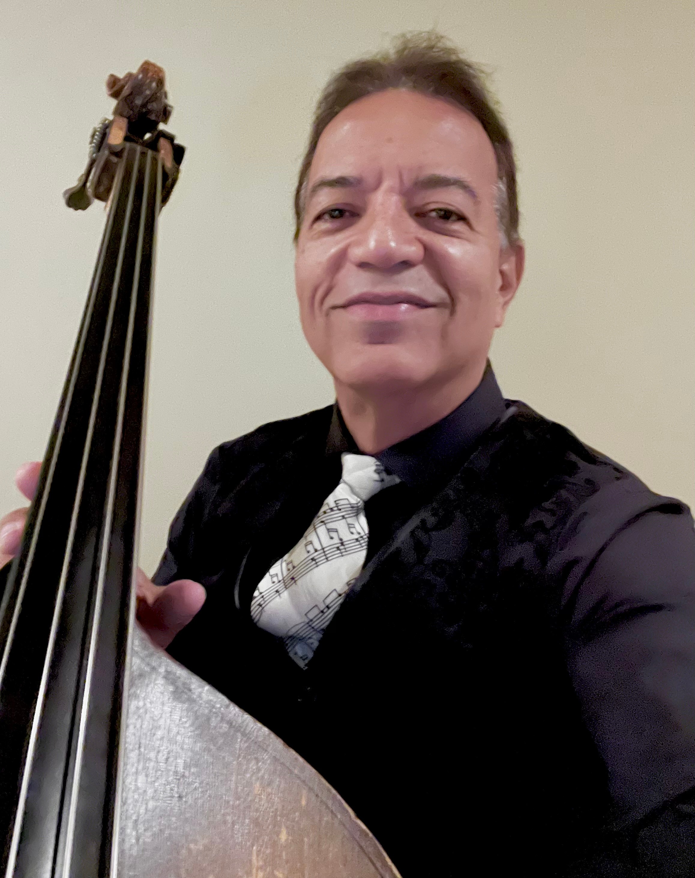 Man smiling holding a string instrument, wearing a black shirt and tie, against a plain background.