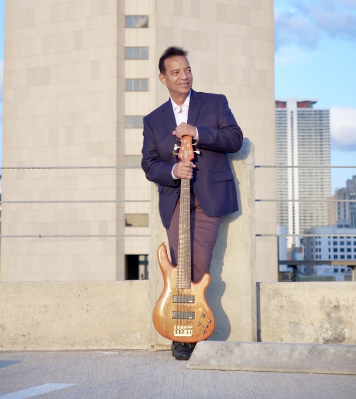 Man in a suit holding a bass guitar on a rooftop with city buildings in the background.