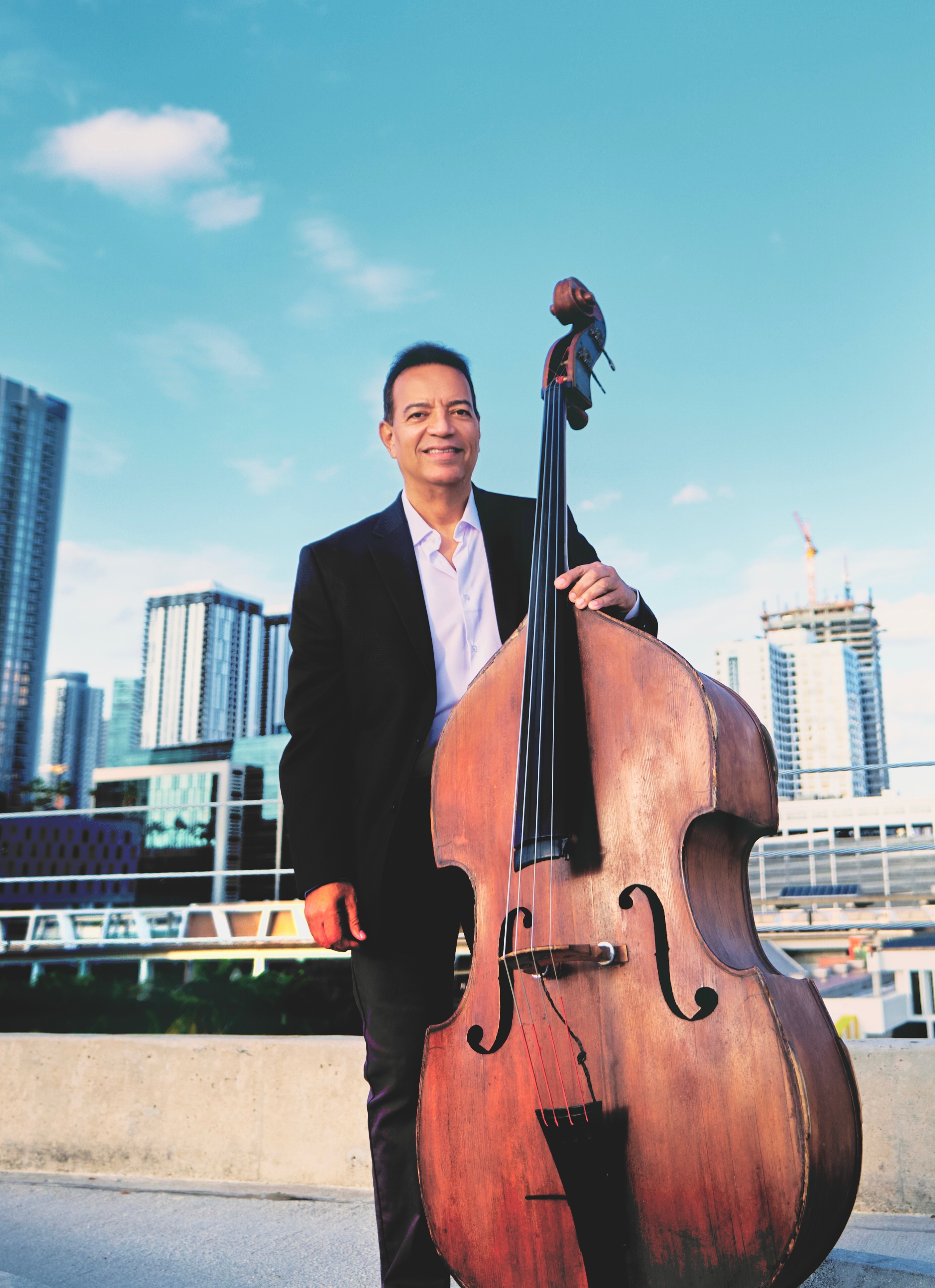 Man in a suit holding a large double bass outdoors with city buildings in the background.