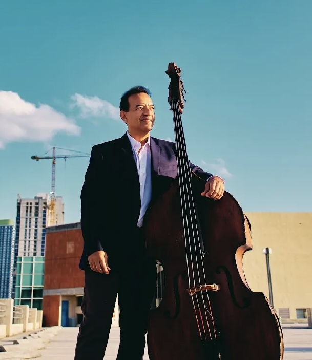 Man in suit standing outdoors next to a large double bass, with a cityscape and blue sky in background.