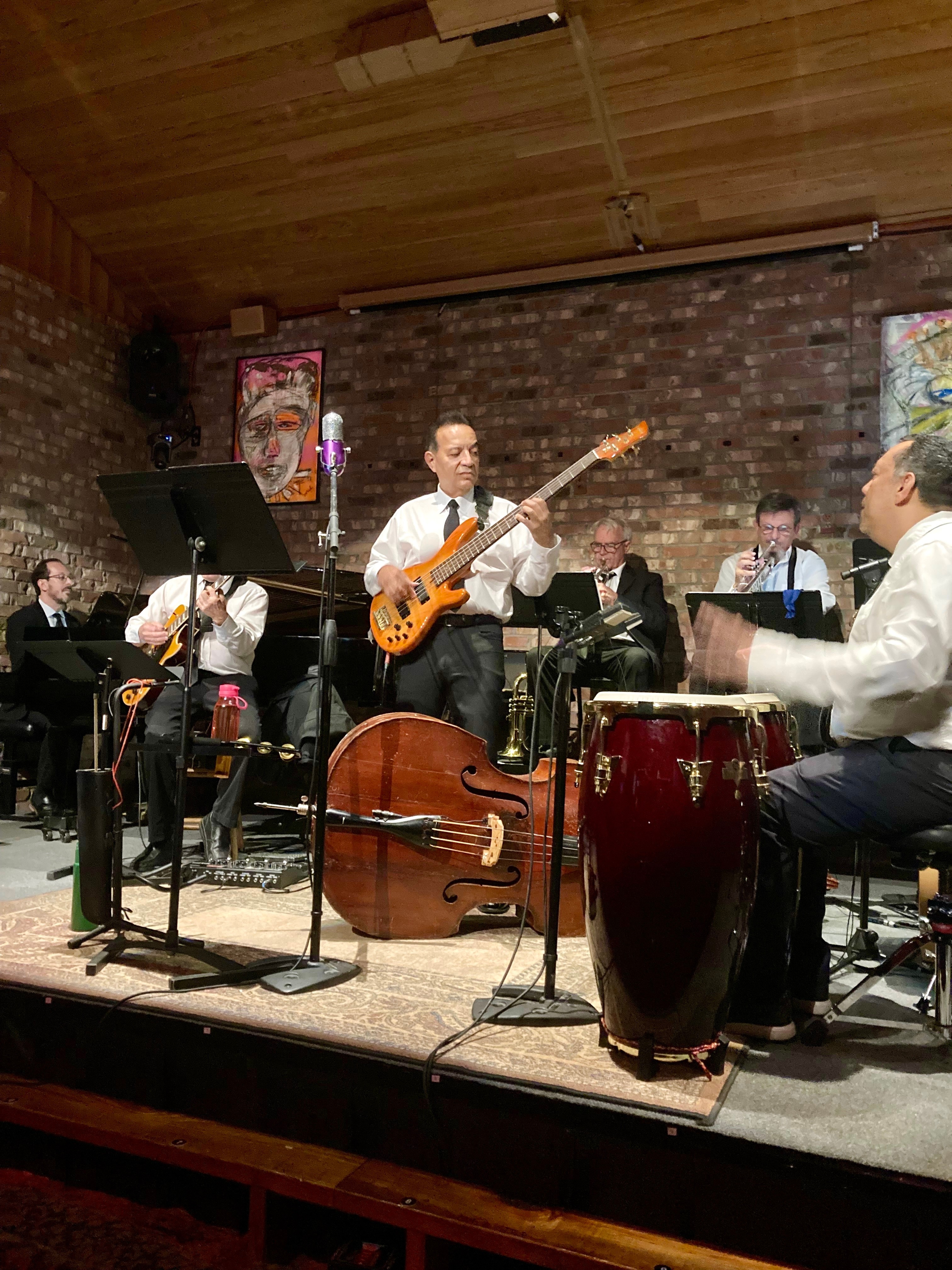 Group of musicians performing on stage with instruments, including a bass guitar, drums, and a piano, in a brick-walled room.