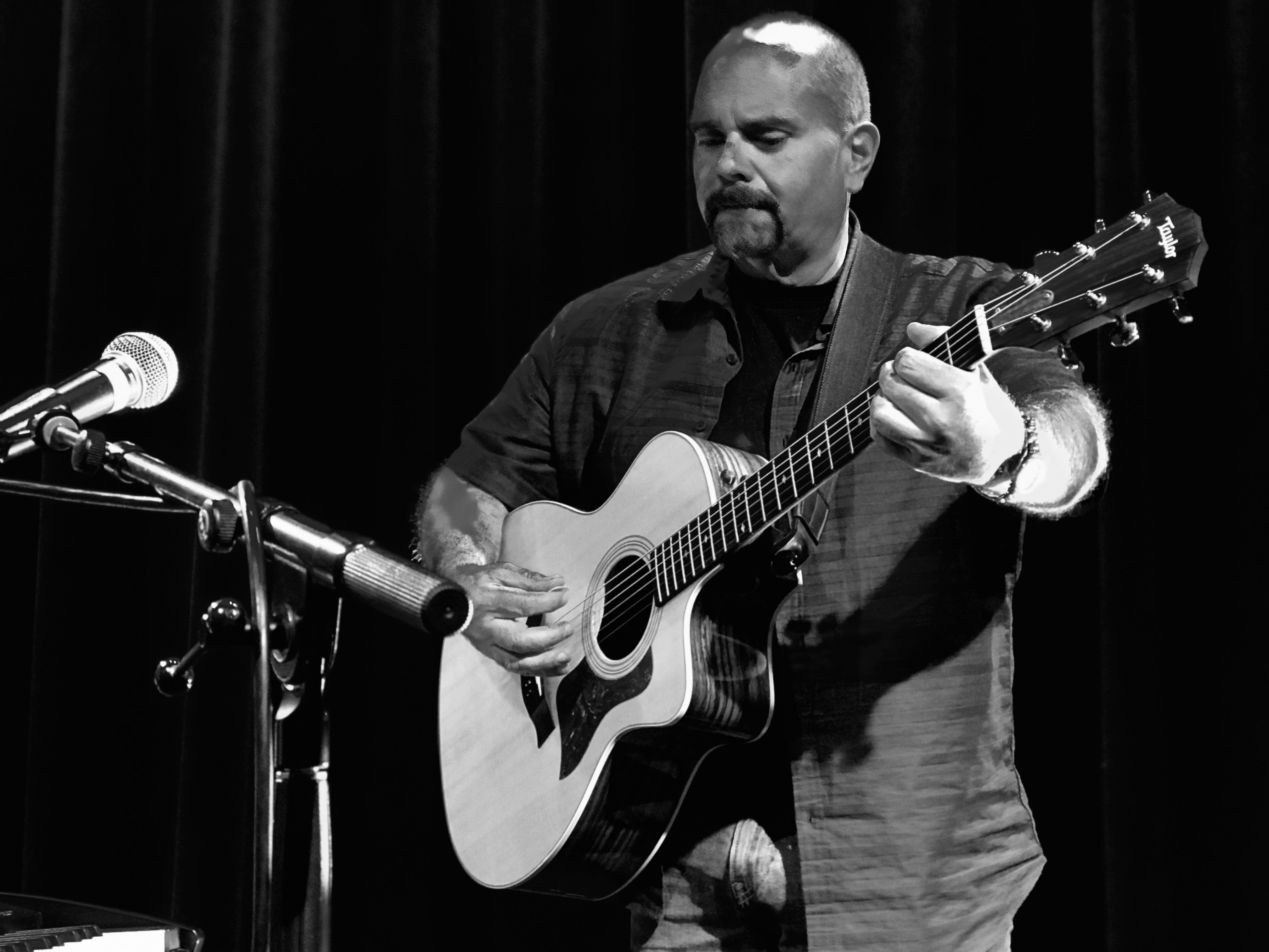 Man playing acoustic guitar on stage with microphone, black curtain background, focused expression