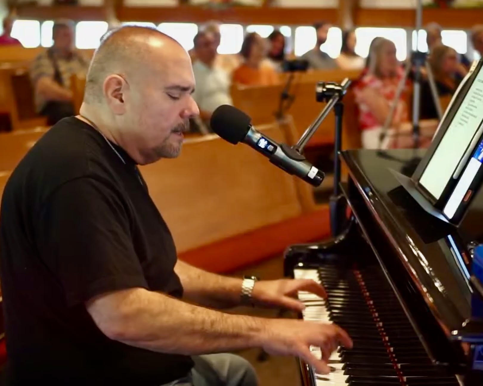 Man playing piano and singing into a microphone in a room with an audience, with sheet music on the piano, in a concert setting.