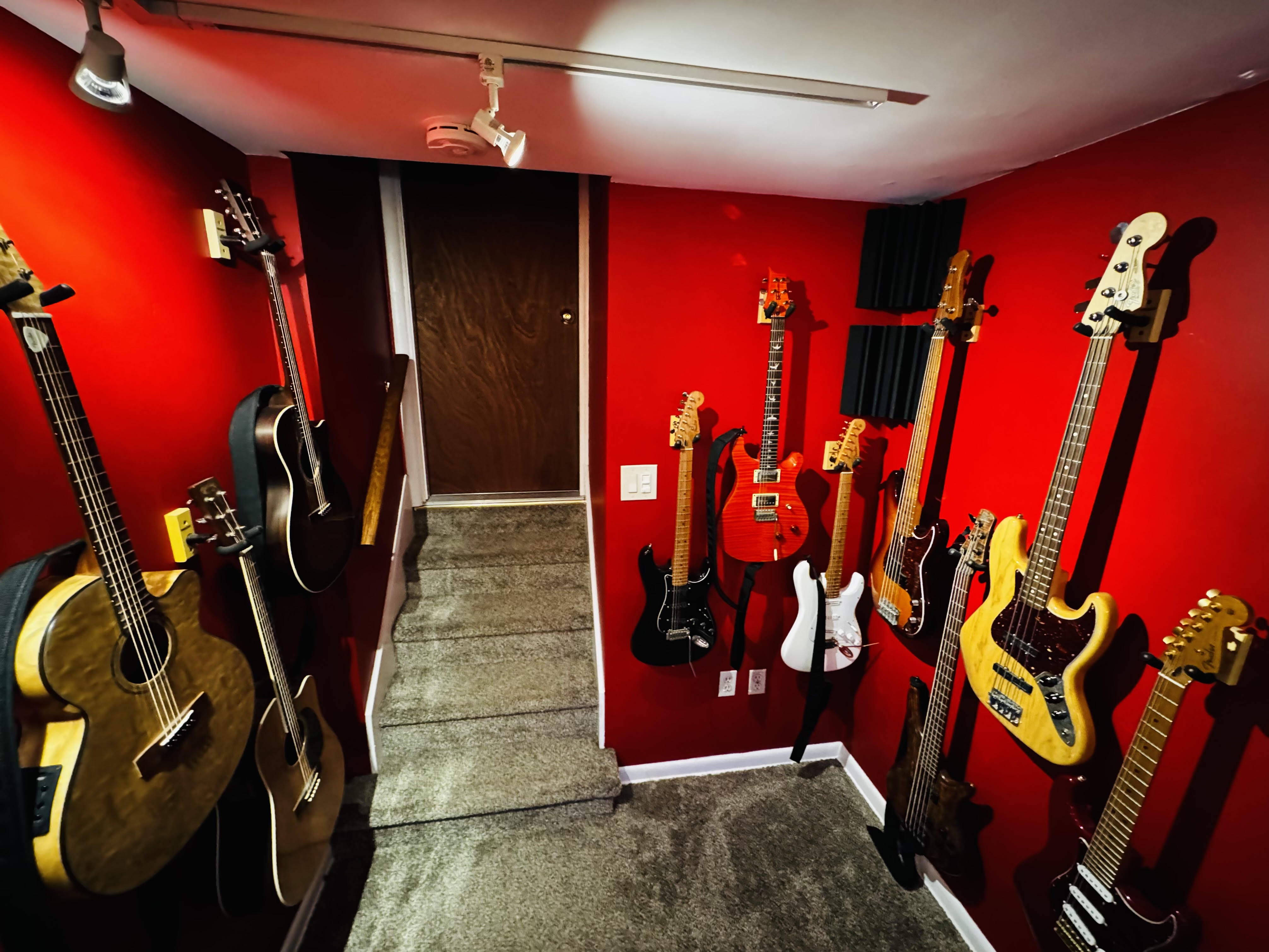 Room with red walls displaying various guitars on stands and wall mounts, door in background, carpeted floor.