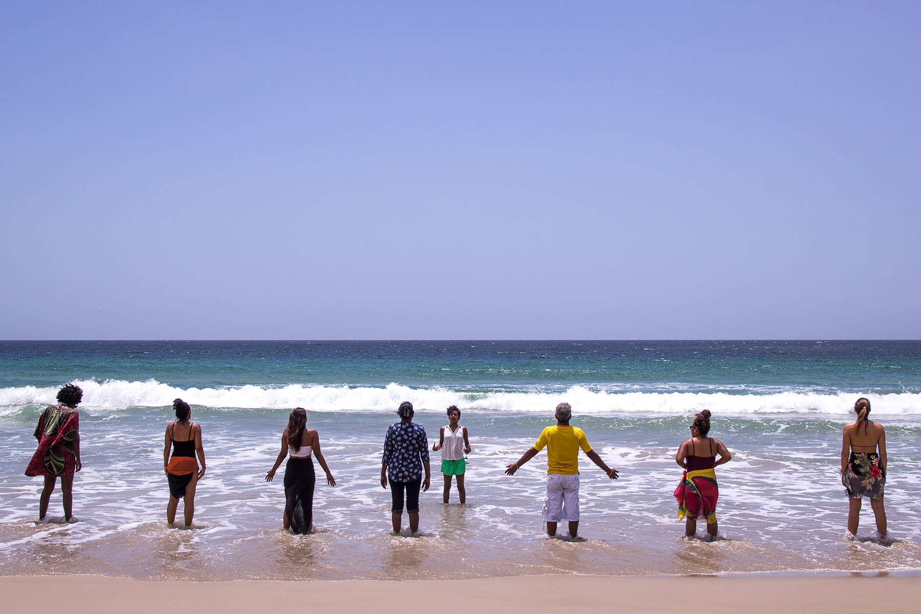 Group of people standing at the shoreline facing the ocean, waves crashing, clear sky, beach scene.