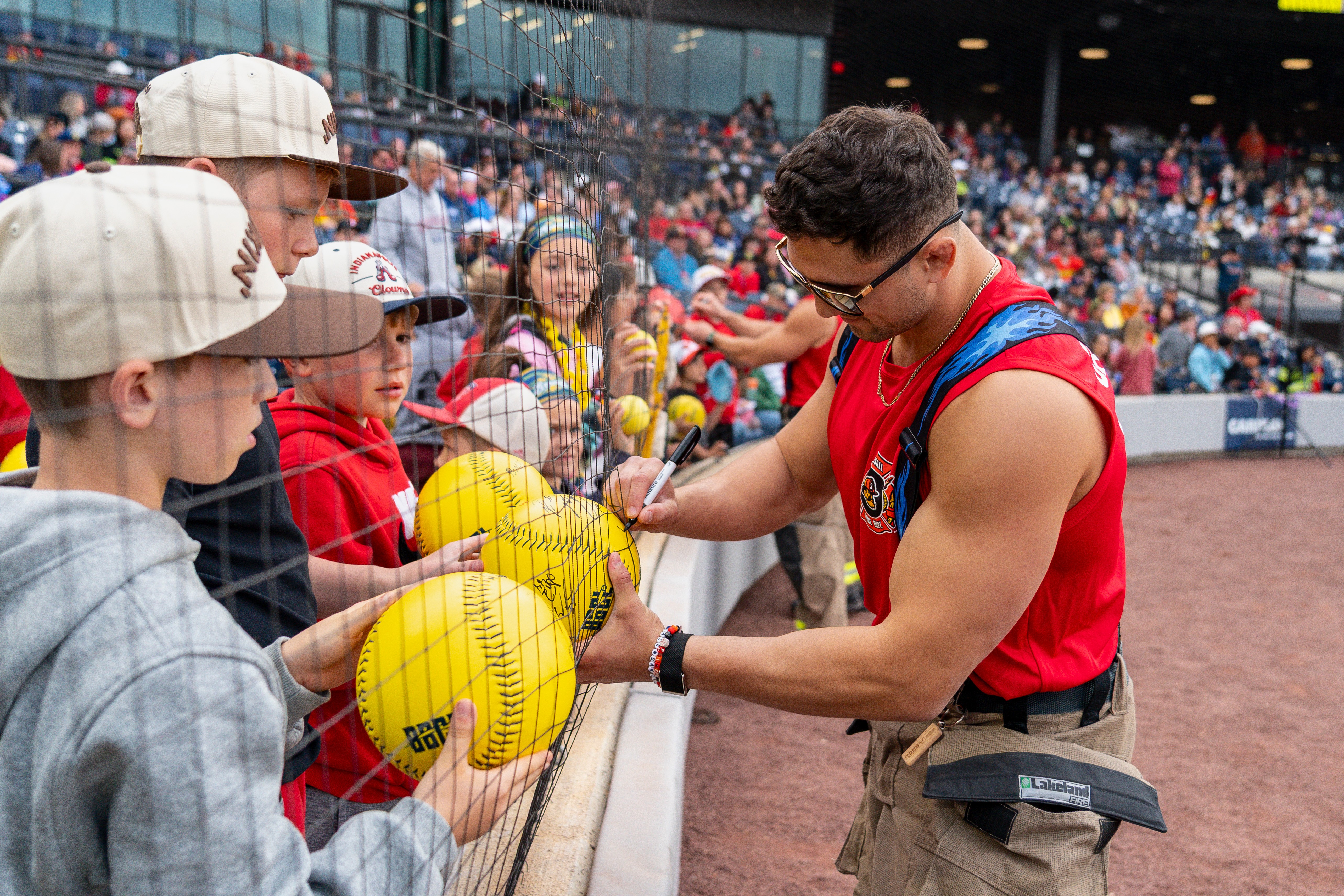 Athlete signing autographs for children holding yellow baseballs at a stadium, with spectators in the background.