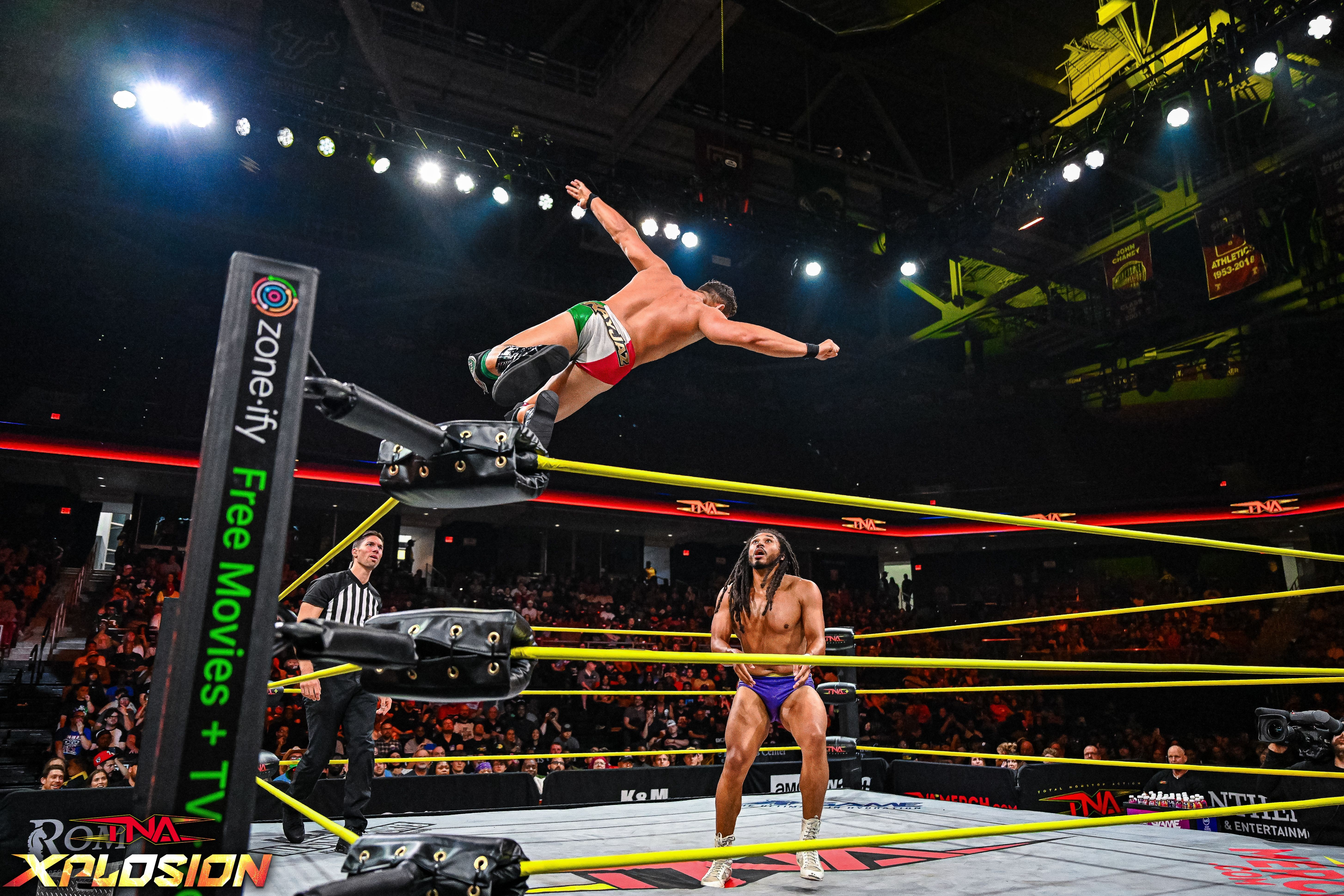 Wrestler in red shorts leaps off the top rope towards another wrestler in the ring, with a crowd watching.