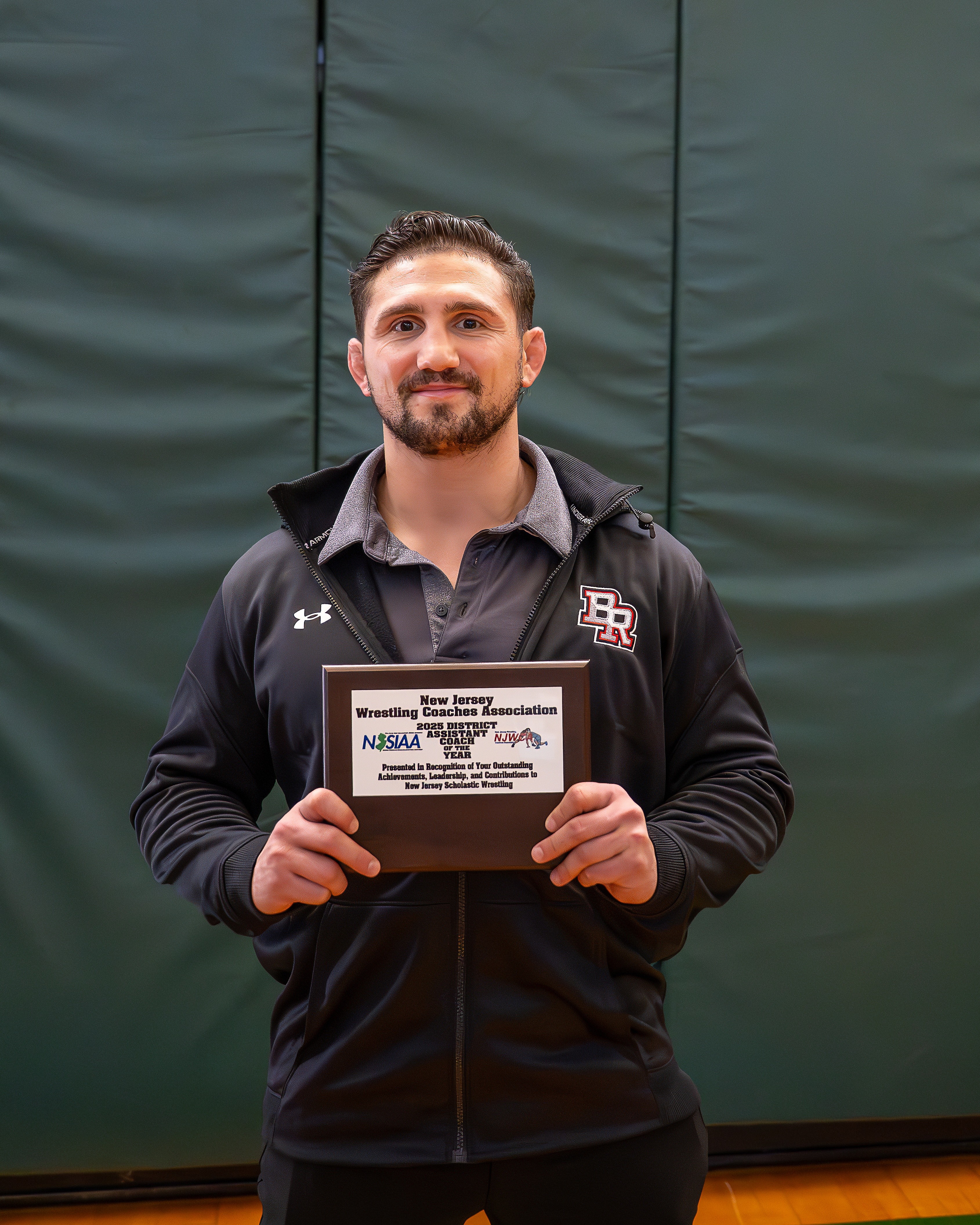 Man holding a certificate, standing indoors in front of a green background, smiling, wearing a black jacket with a logo.