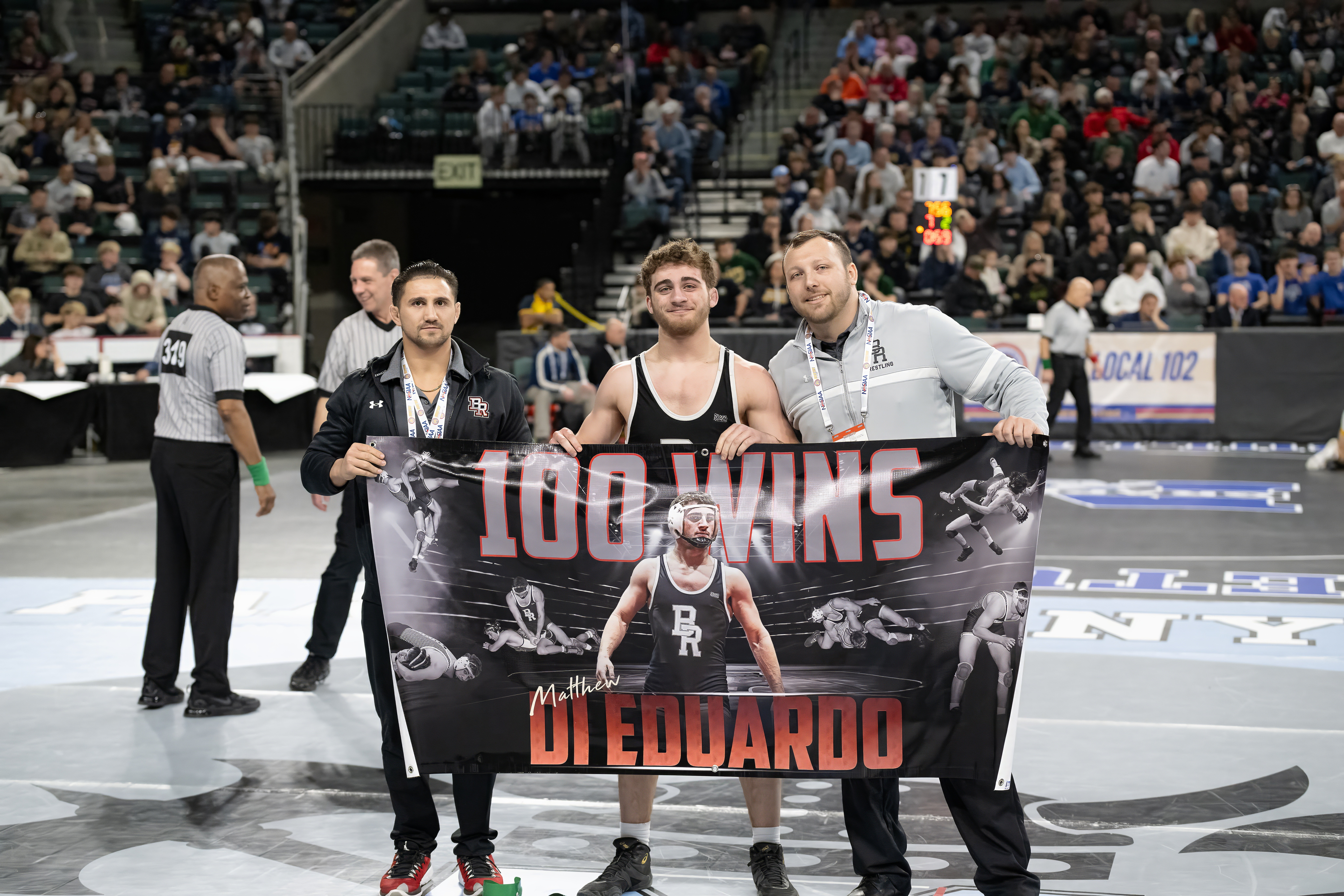 Three men holding a banner with a basketball player, in a sports arena with spectators and a basketball court.