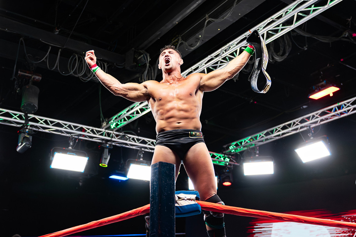 Wrestler stands on ring with arms raised, holding a championship belt, in a wrestling arena with bright lights and rigging.