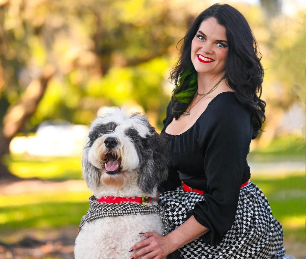Woman with dark hair and red lipstick smiling outdoors with a black and white dog, trees in background.