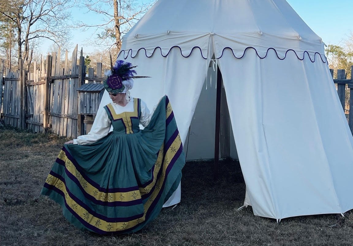 Person in historical costume standing outside a white tent, holding the skirt of their dress, with a wooden fence and trees in background.