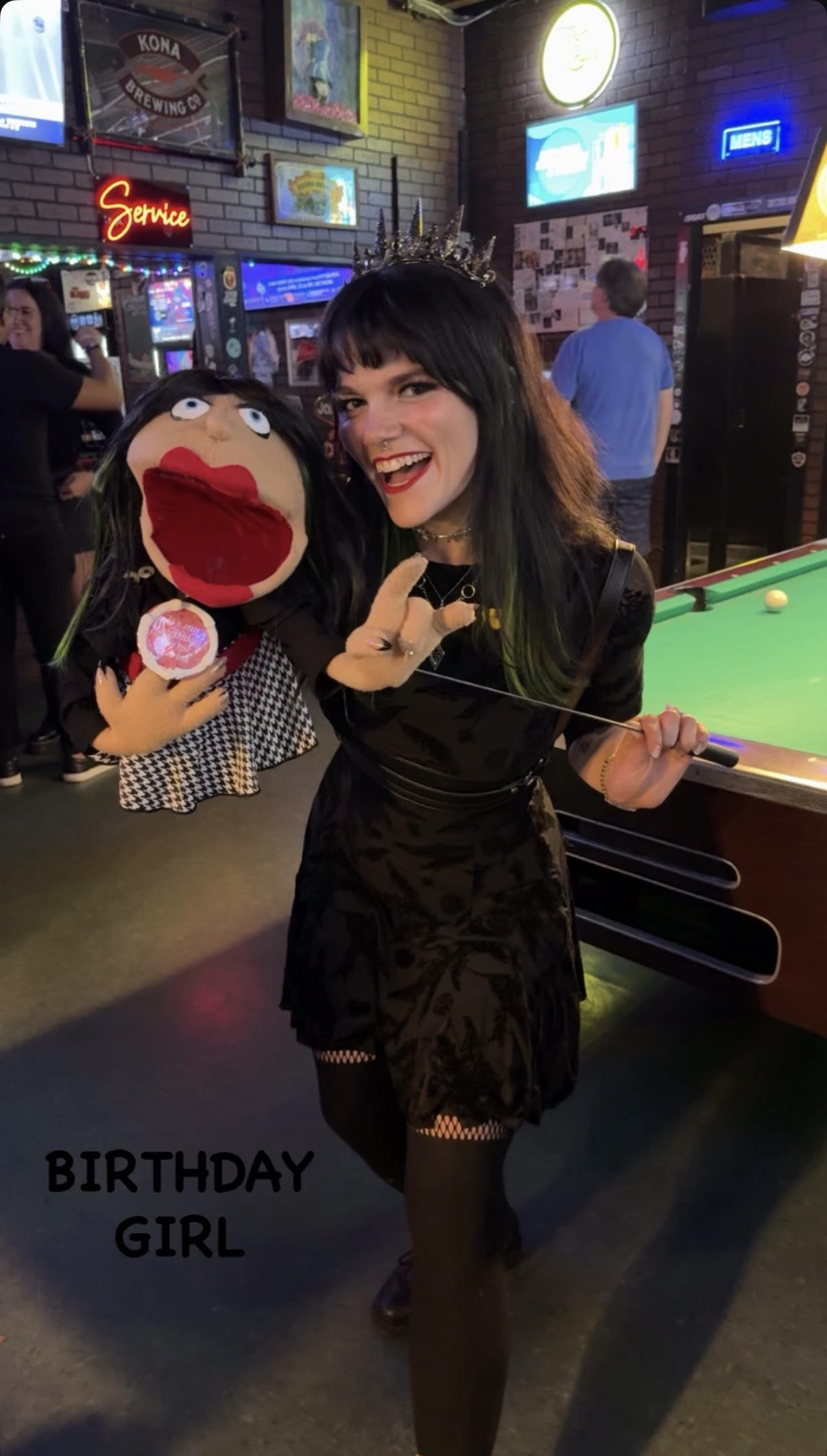 Young woman with dark hair smiling and holding a puppet in a bar with pool tables and neon signs.