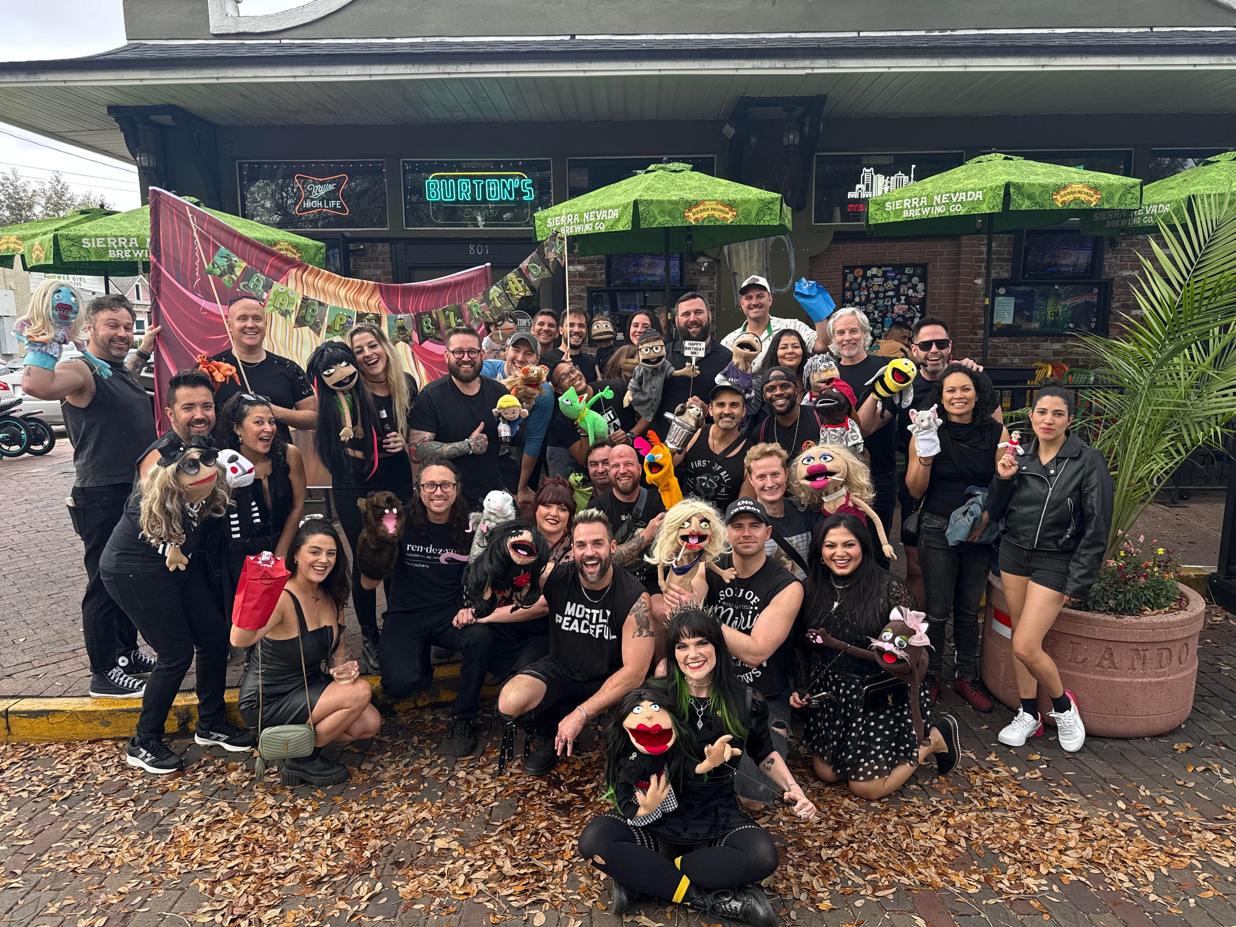Group of people gathered outside a store, some holding drinks, with umbrellas and greenery, on a rainy day.
