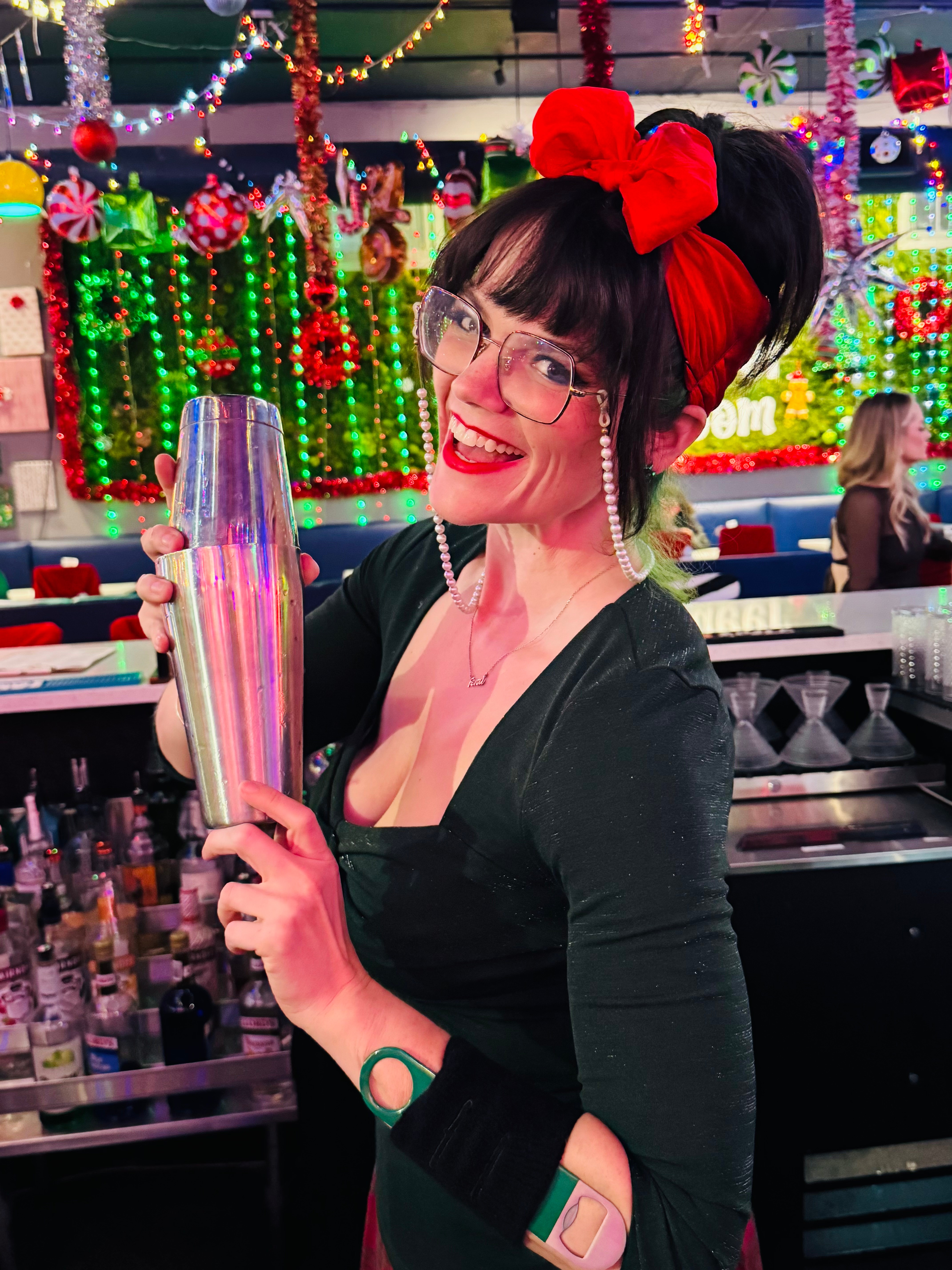 Woman with glasses, red lipstick, and a large red bow in her hair, holding a colorful bottle, in a decorated festive setting.