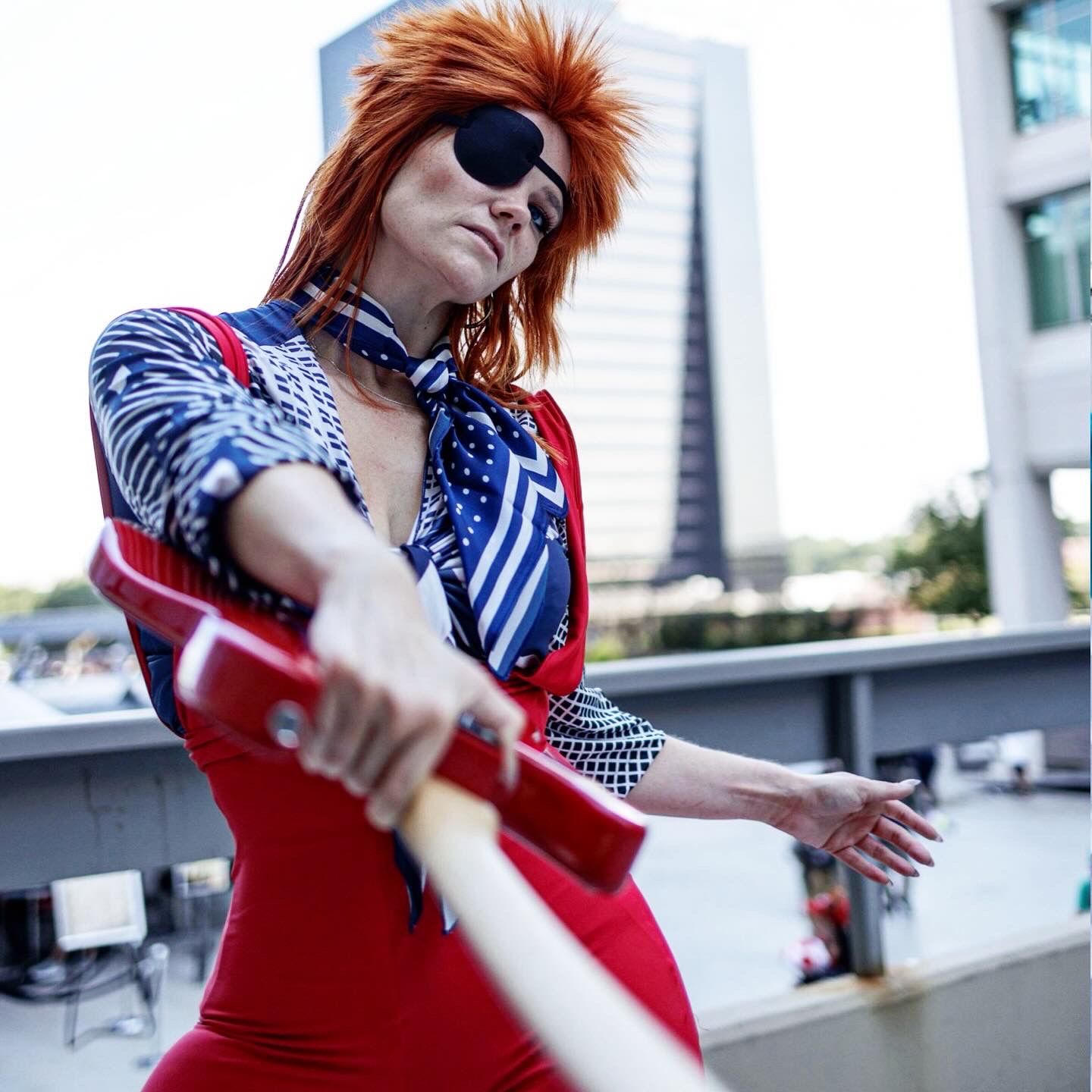 Person with red hair, sunglasses, and patterned clothing holding a red and white object outdoors in an urban setting.