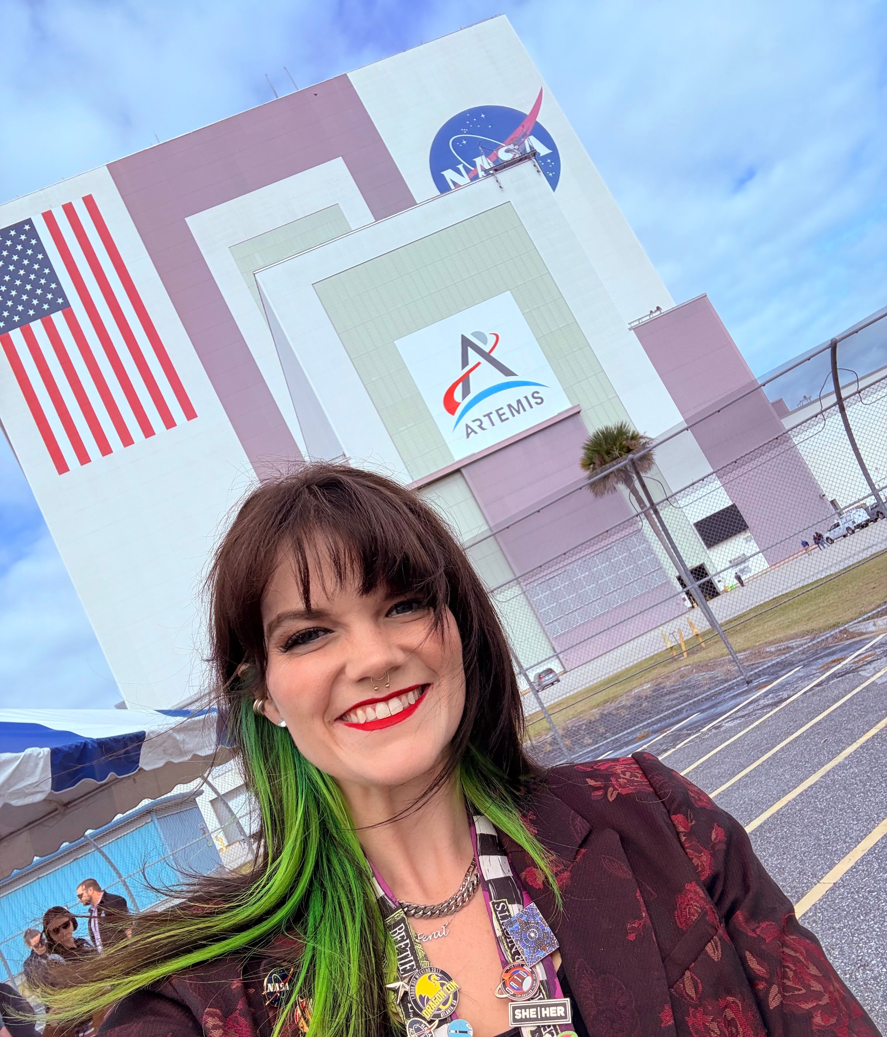 Smiling woman with green hair and red lipstick in front of NASA and Artemis logos, parking lot, and a building, sky background.