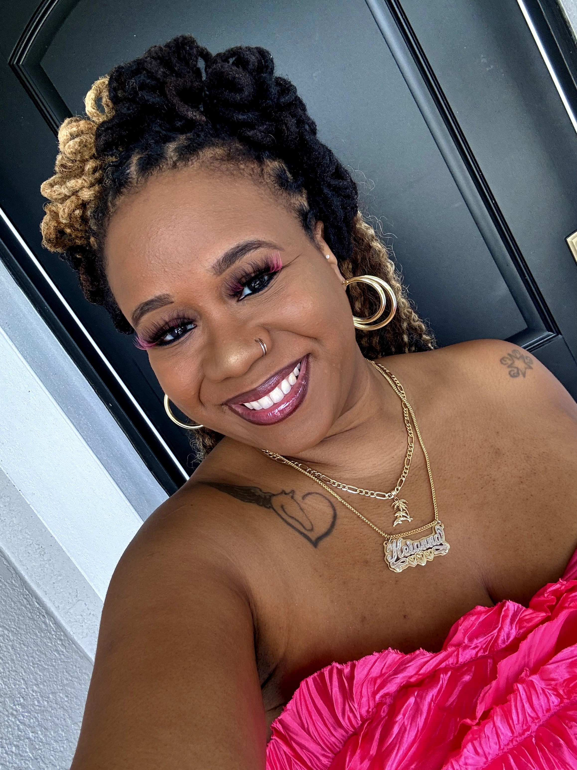Smiling woman with styled hair, gold jewelry, and a pink top, taking a selfie indoors.