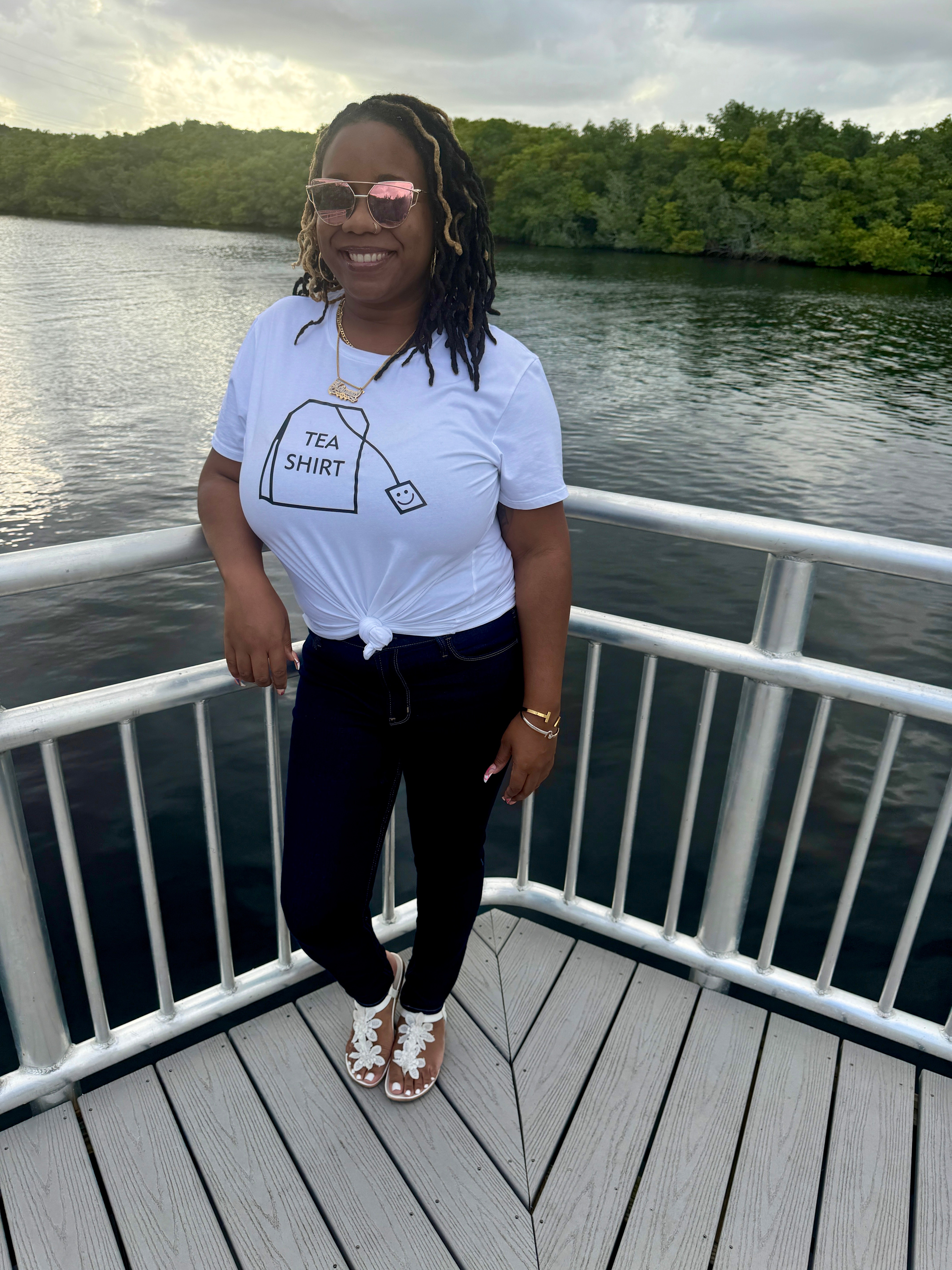 Woman standing on boat deck near water, smiling, wearing sunglasses, white T-shirt, black pants, and sandals.