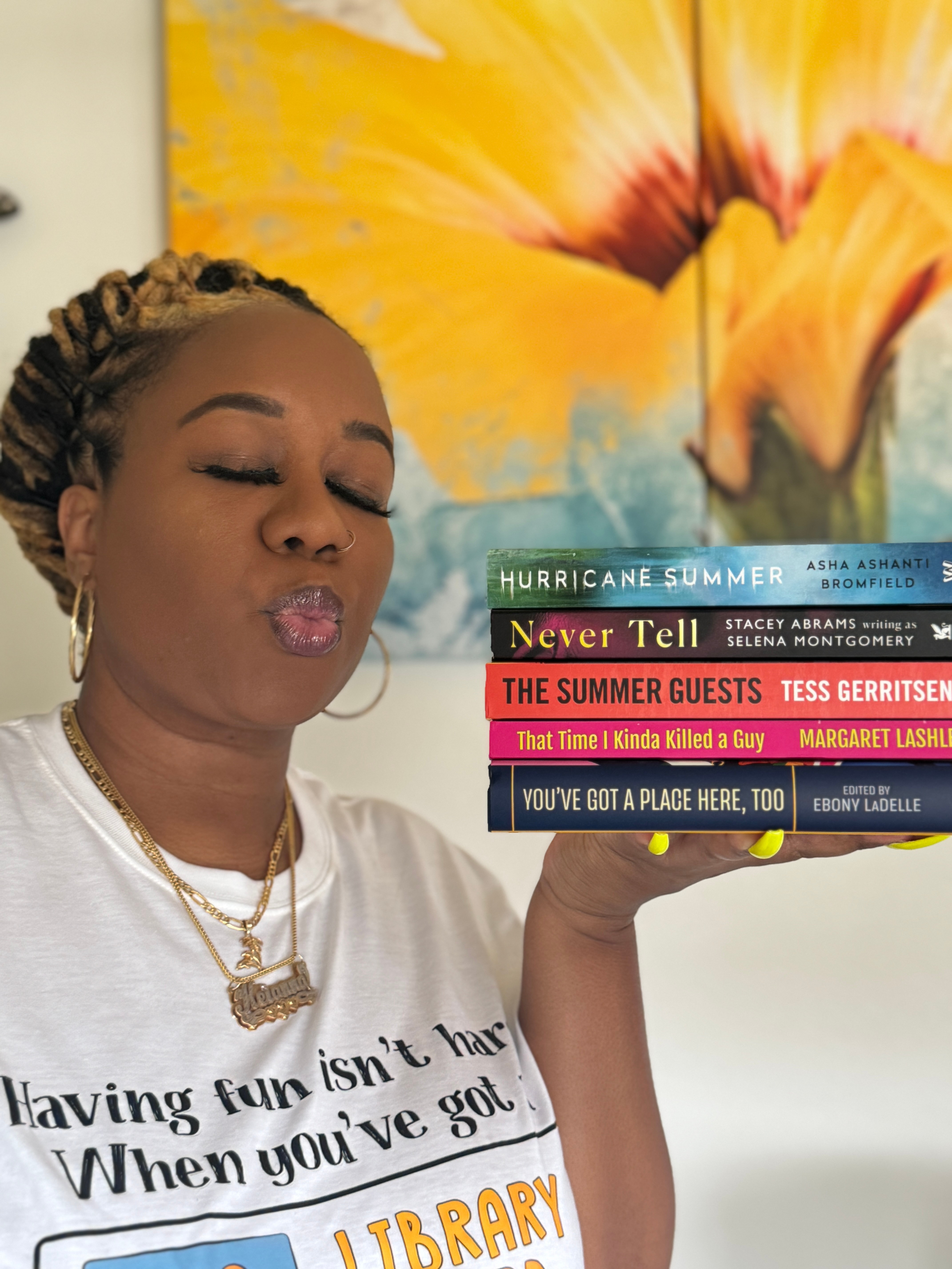 A woman with braided hair and jewelry holds a stack of colorful books, standing in front of a yellow floral background.