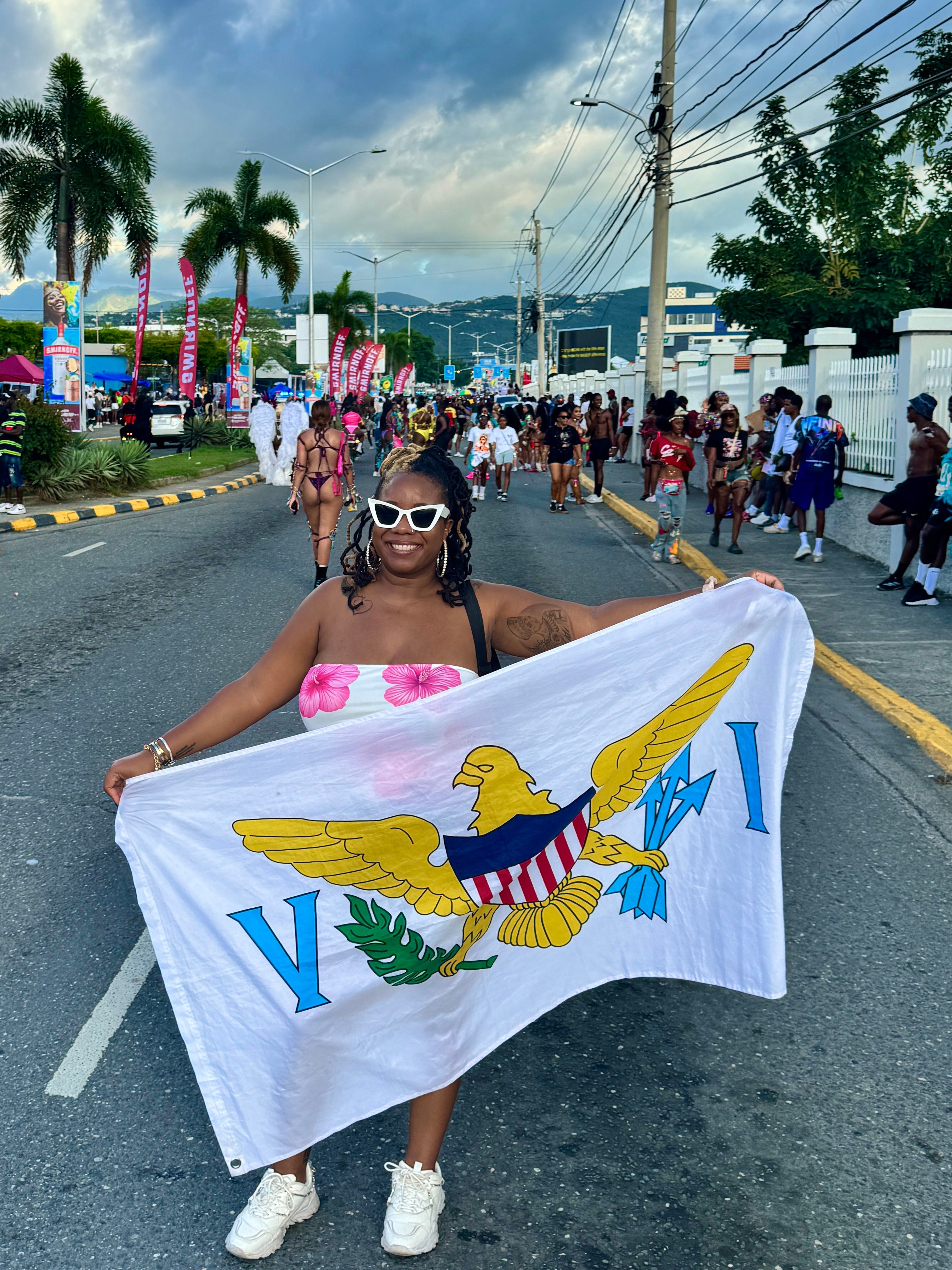 Woman holding a flag with a bird emblem, wearing sunglasses and a pink top, on a street with people and palm trees.