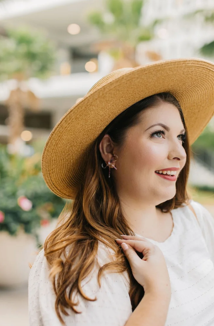 Woman wearing a wide-brimmed hat outdoors, smiling, with blurred greenery and buildings in background.