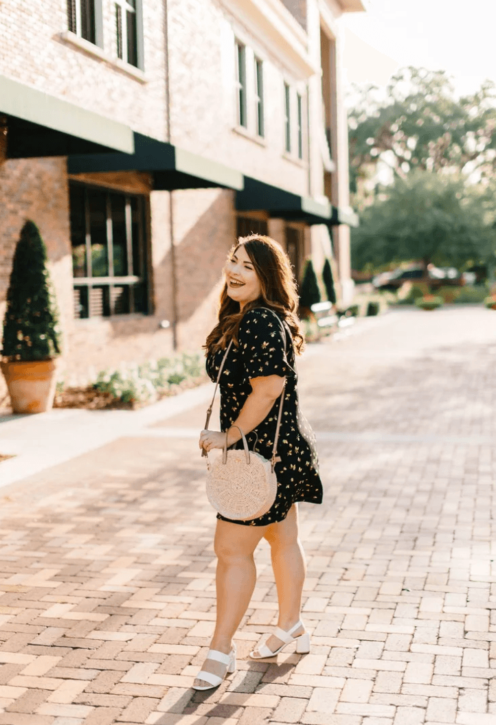 Young woman in a black dress with white polka dots walking on a brick sidewalk, holding a white bag, smiling.