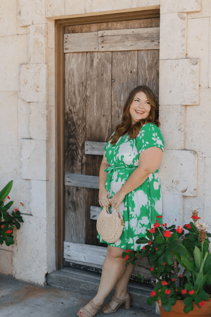 Woman in green and white dress standing in front of a wooden door, smiling, with potted plants nearby.