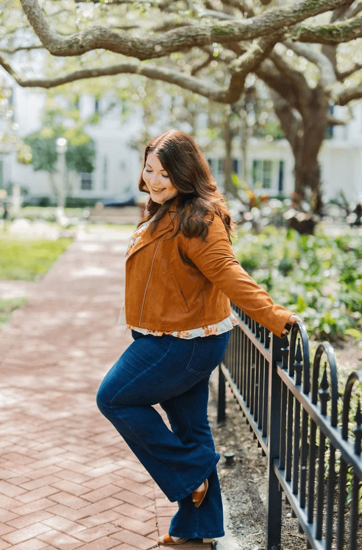 Woman with long hair leaning on a black fence in a park, smiling, wearing a brown jacket and blue jeans.
