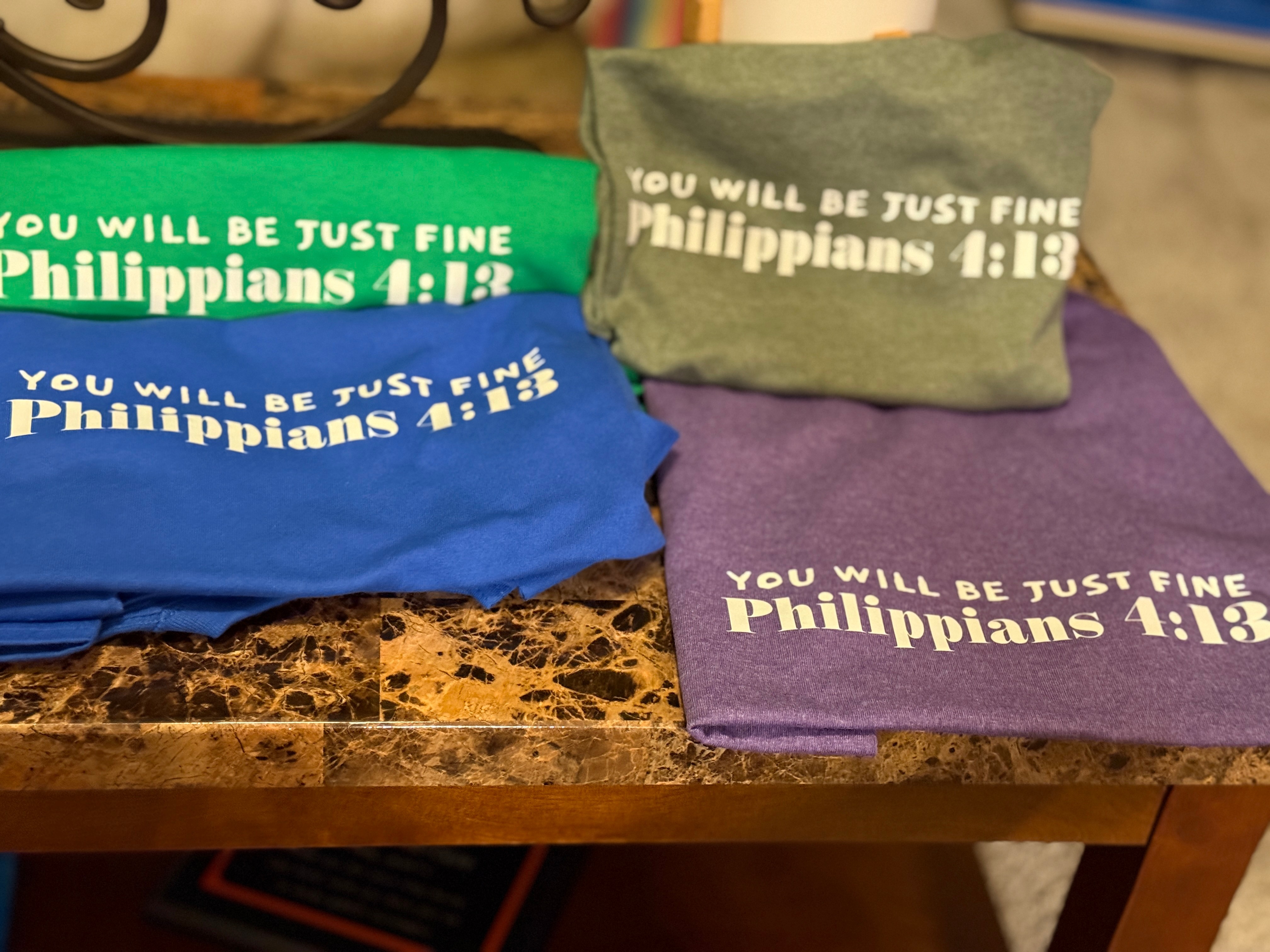 Three folded T-shirts in green, blue, and purple with white text on a marble surface.