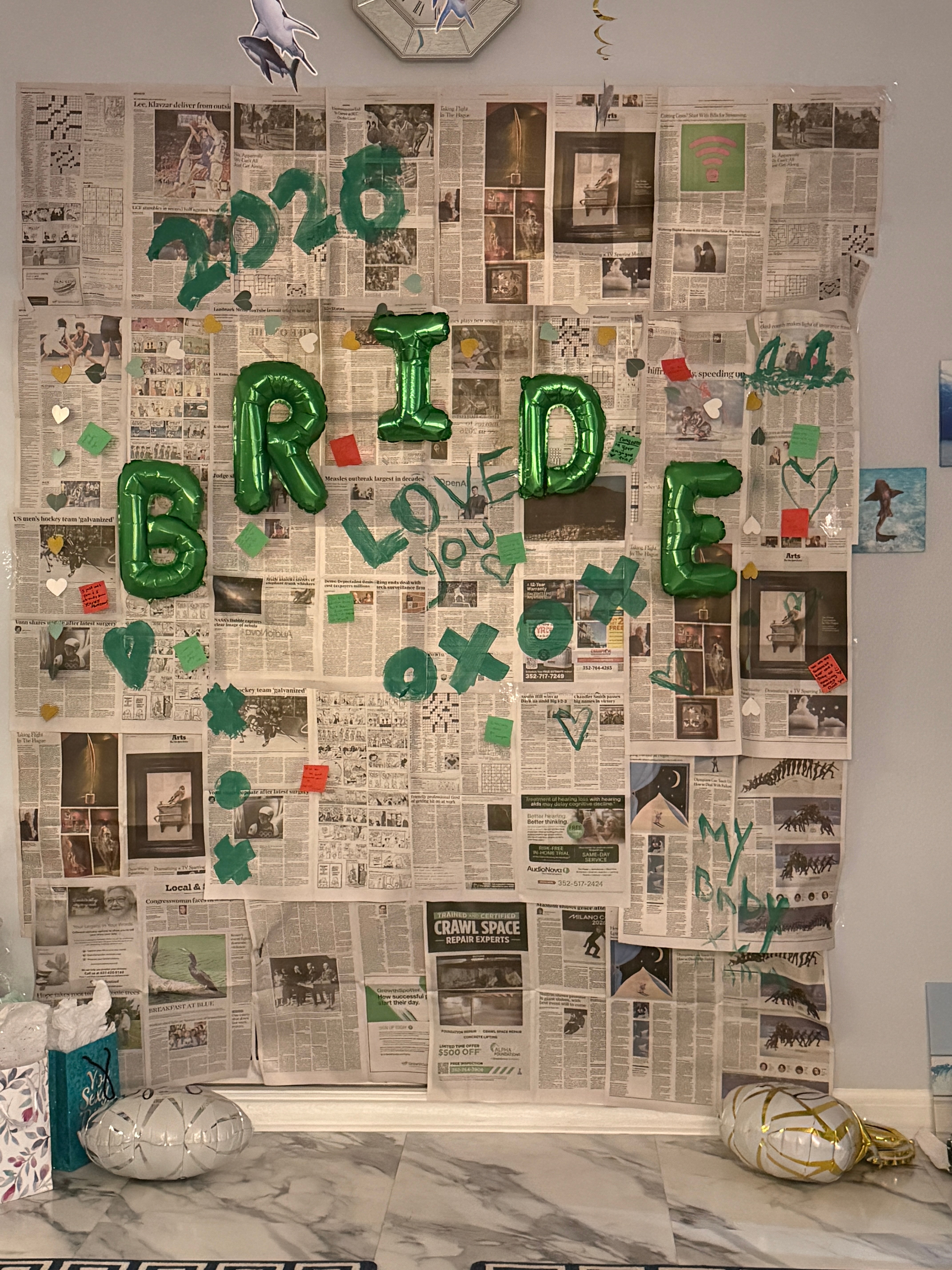 Wall decorated with newspaper clippings, green balloons spelling 'BRIDE' and 'LOVE', and various small decorations.