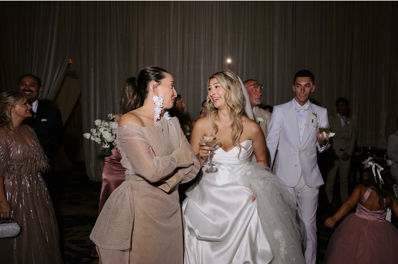 Group of people at a formal event, two women in conversation, one in a white dress, others in elegant attire, dim lighting.