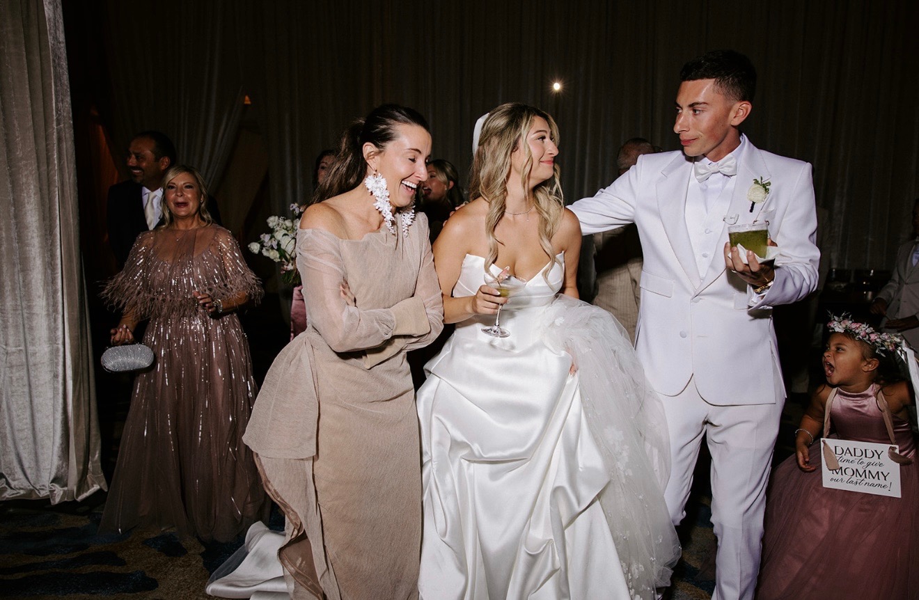 Three people dancing at a wedding reception, with a woman in a white dress in the center, a man in a white suit, and another woman in a beige dress, smiling and holding drinks.