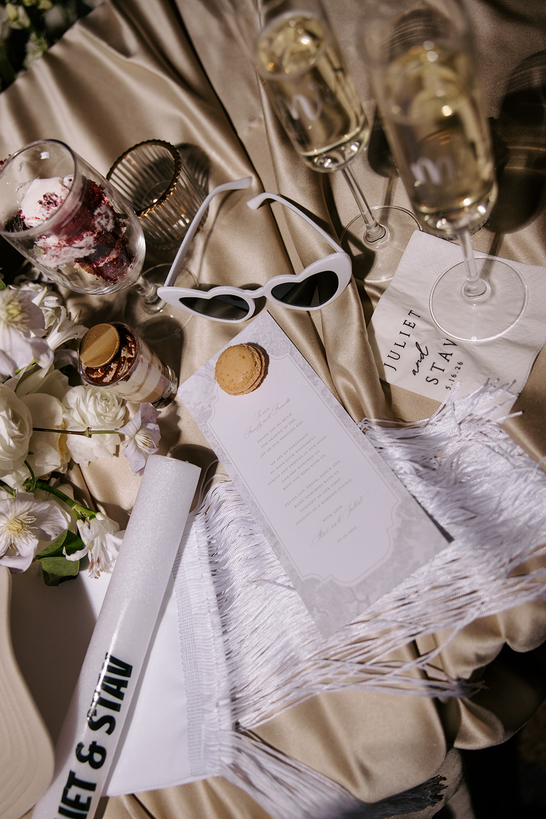 Table setting with glasses, flowers, sunglasses, and printed materials on a satin tablecloth.