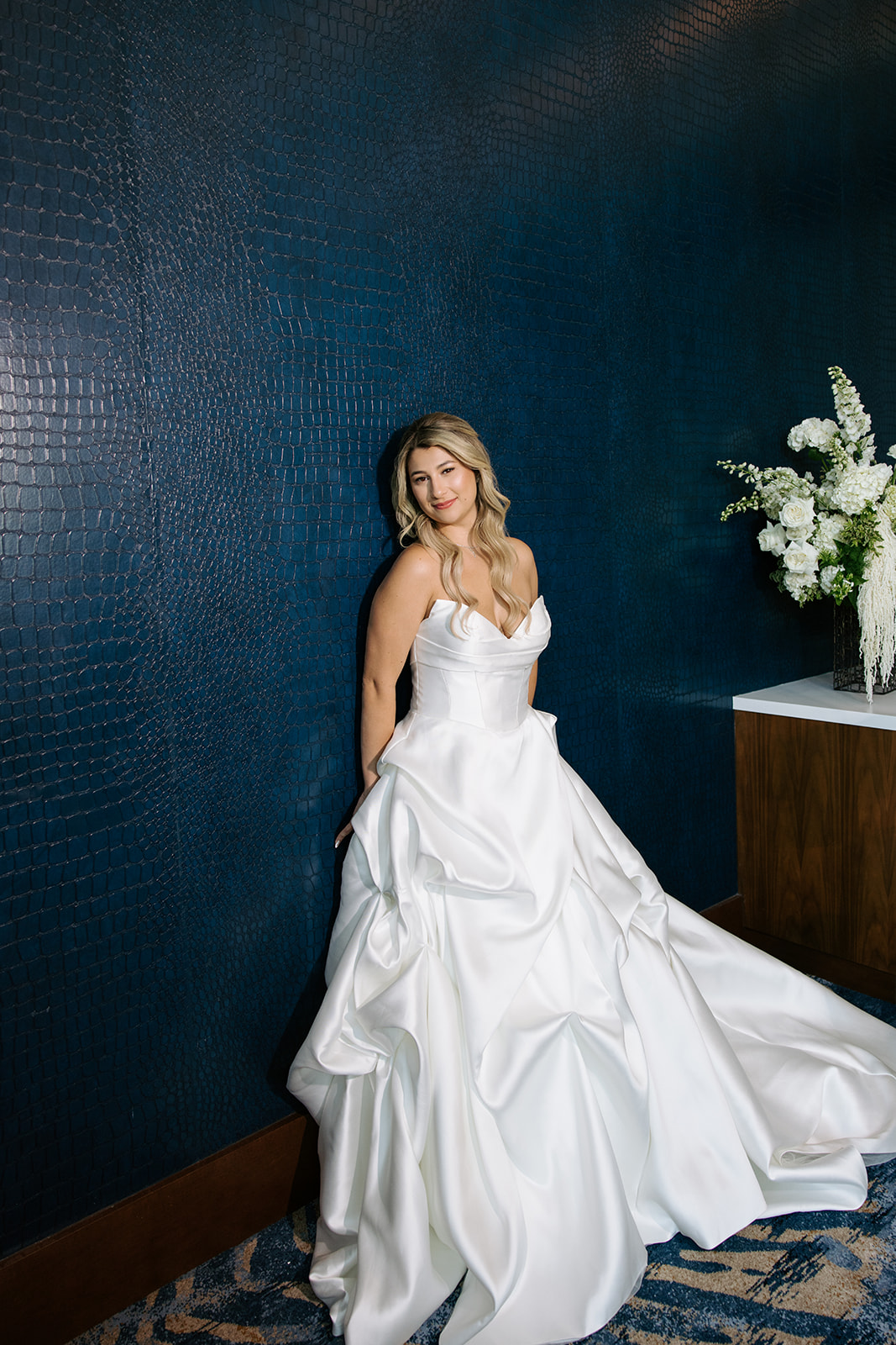 Woman in wedding dress leaning against a dark blue wall with a floral arrangement nearby.