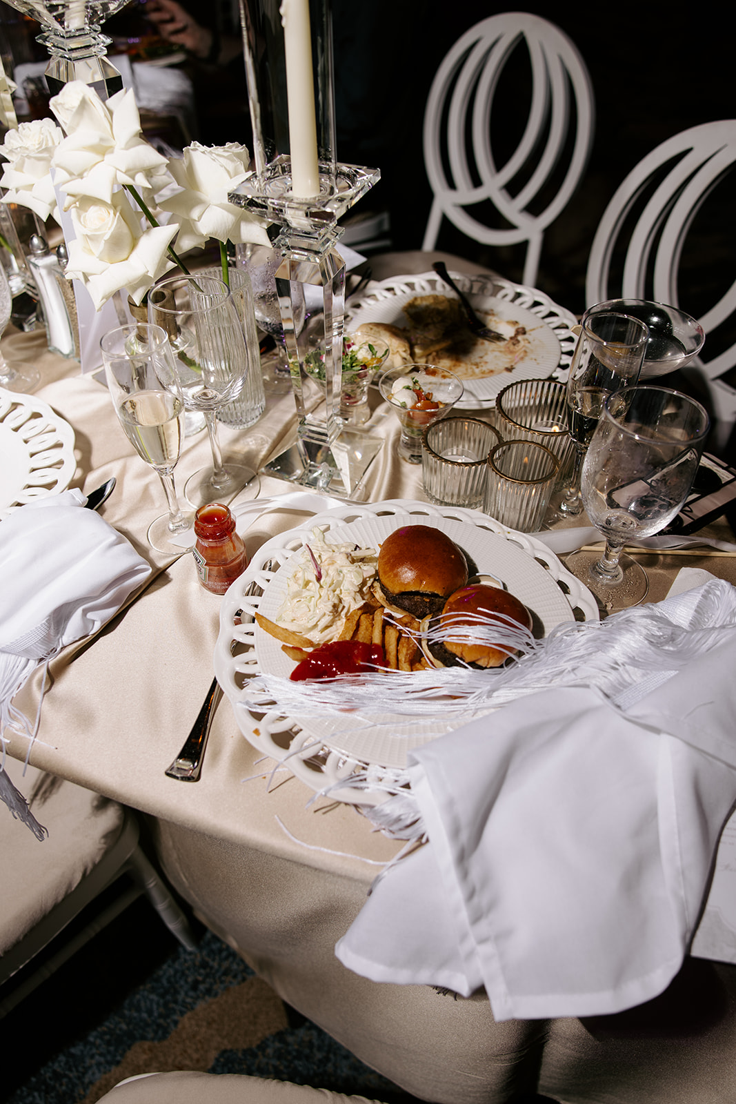 Table with plates, glasses, flowers, and a partially eaten meal, including a burger and fries, with a white tablecloth and decorative elements.