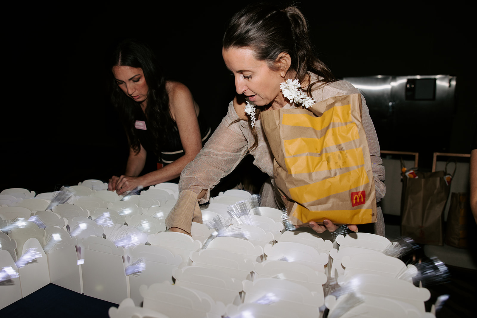 Two women work with illuminated white boxes on a dark surface, one wearing a yellow apron and the other in black.