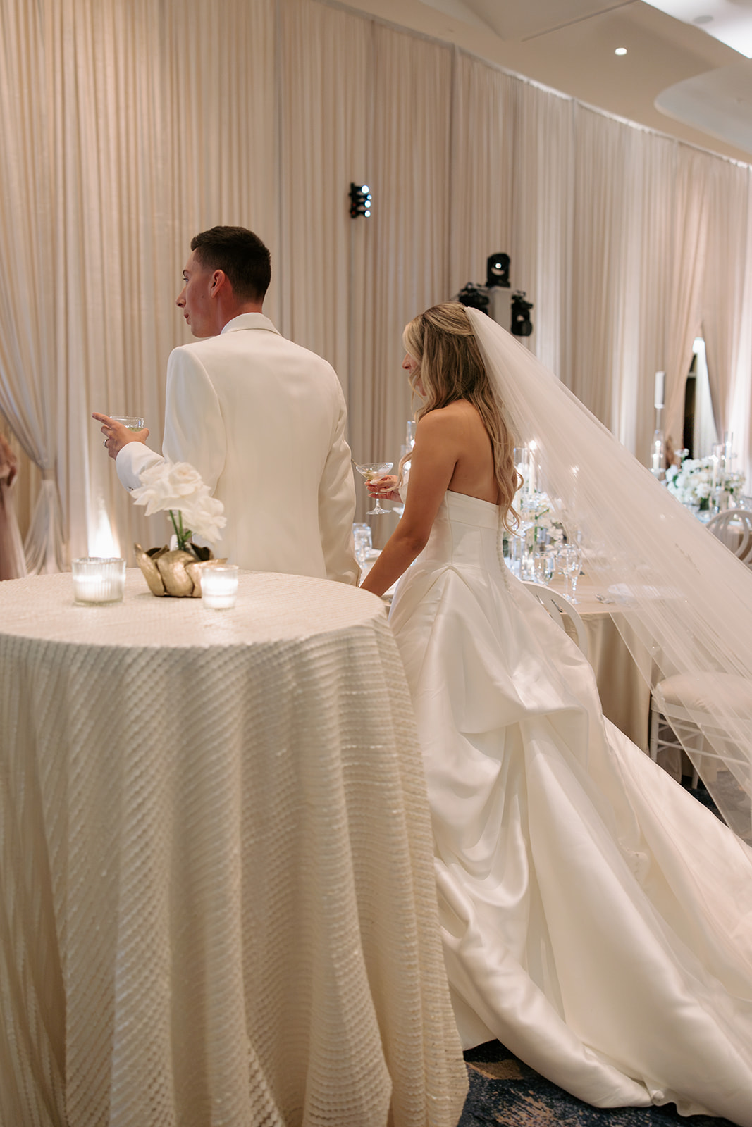 Bride and groom standing at a wedding reception table, with the bride in a wedding gown and veil, and the groom in a suit, in a decorated venue.