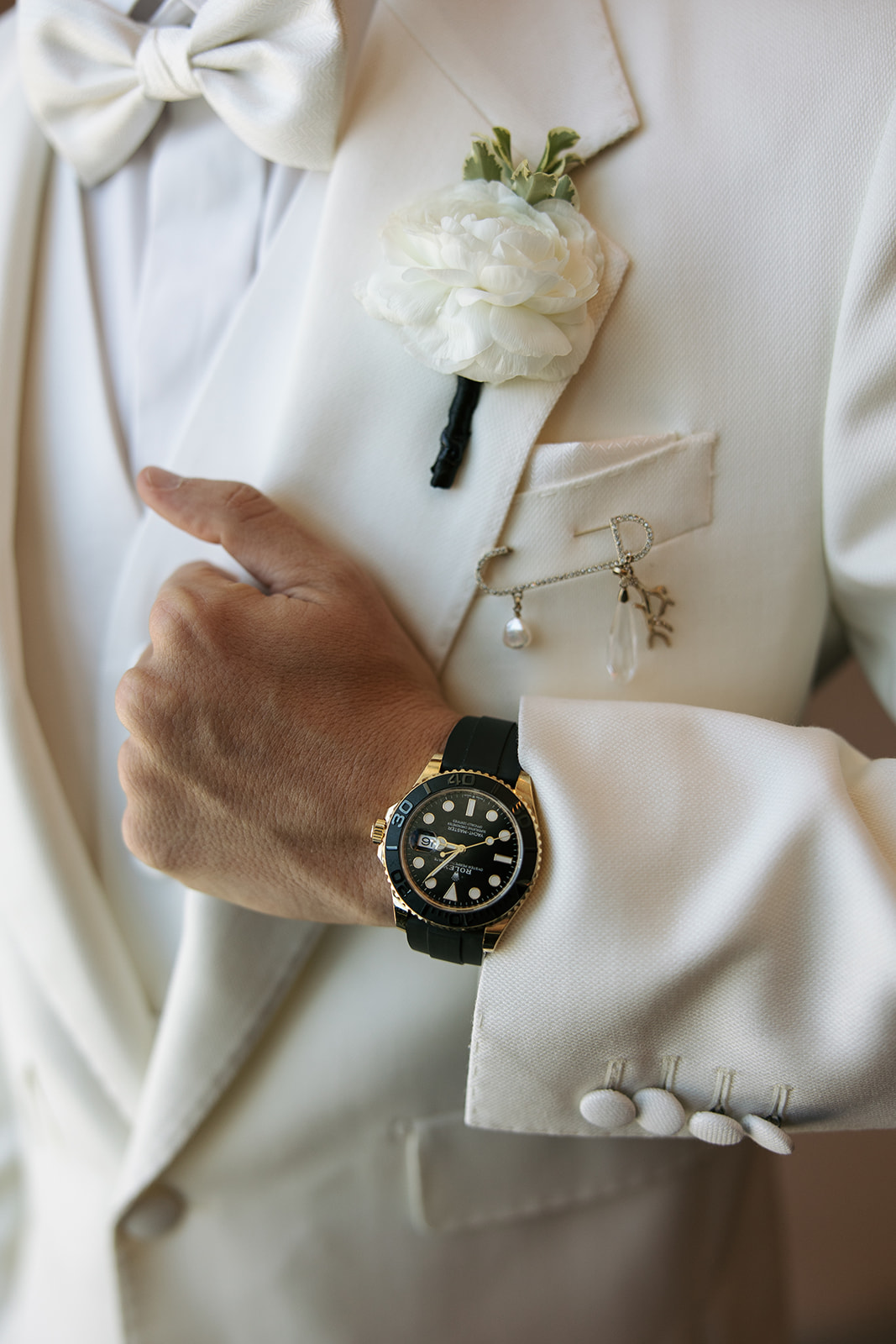 Close-up of a person in a white suit with a boutonniere, watch, and accessories, holding their hand near their chest.