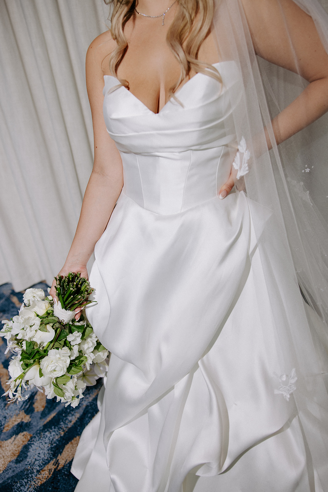 Woman in wedding dress holding bouquet, with veil and floral details, standing indoors.