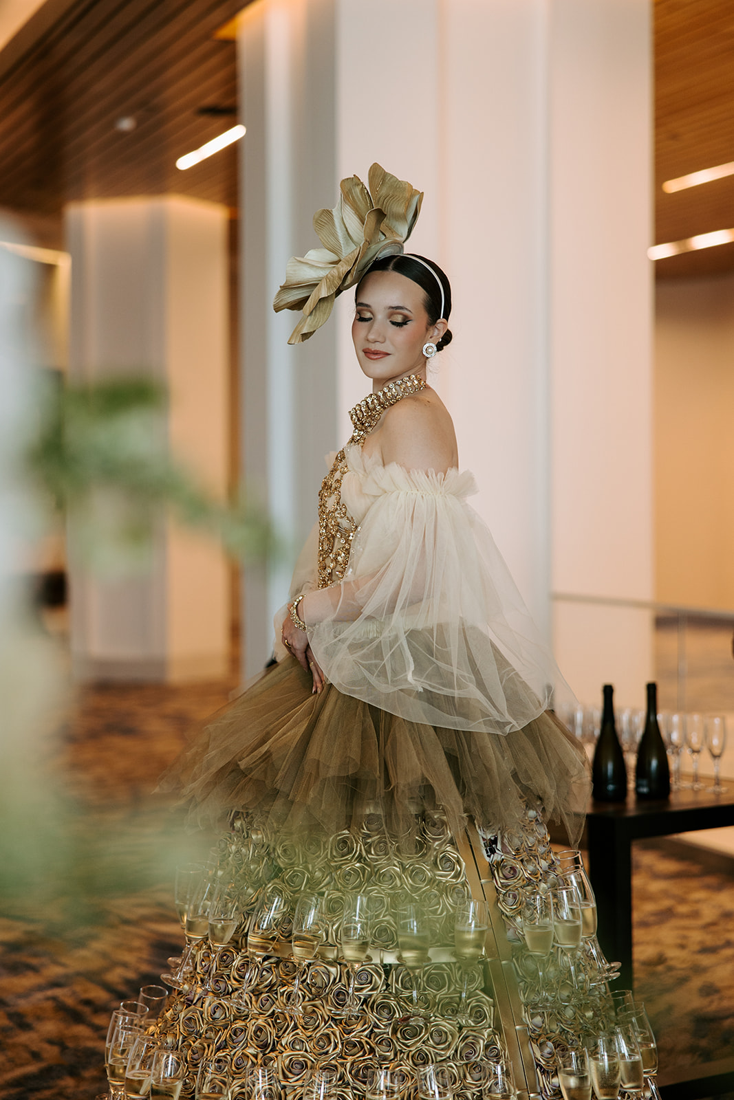 Woman in elaborate gown and headpiece standing indoors with modern decor, soft lighting, and bottles on a table.