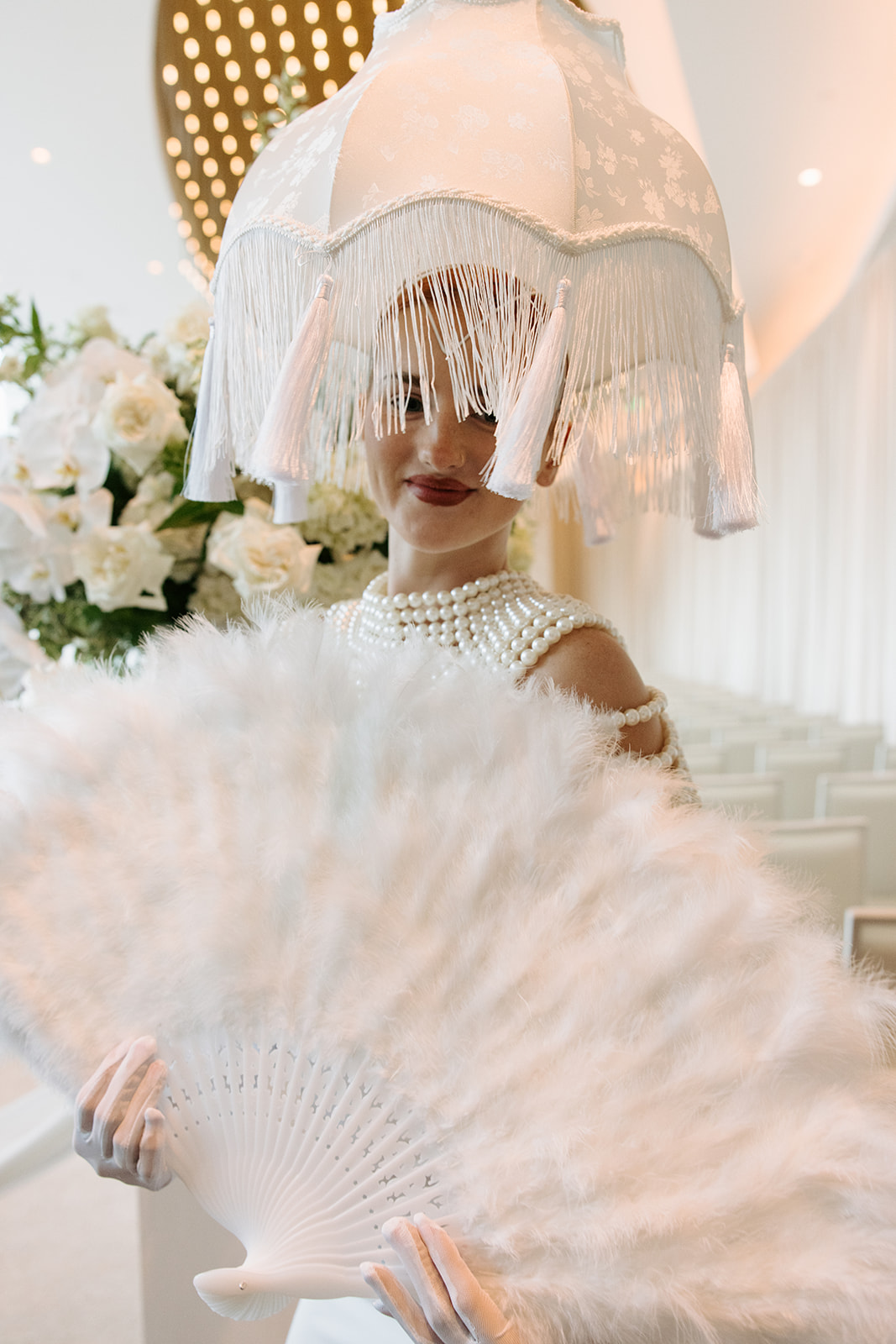 Woman in vintage dress with large hat and pearl necklace holding a feather fan, standing indoors with floral decor.