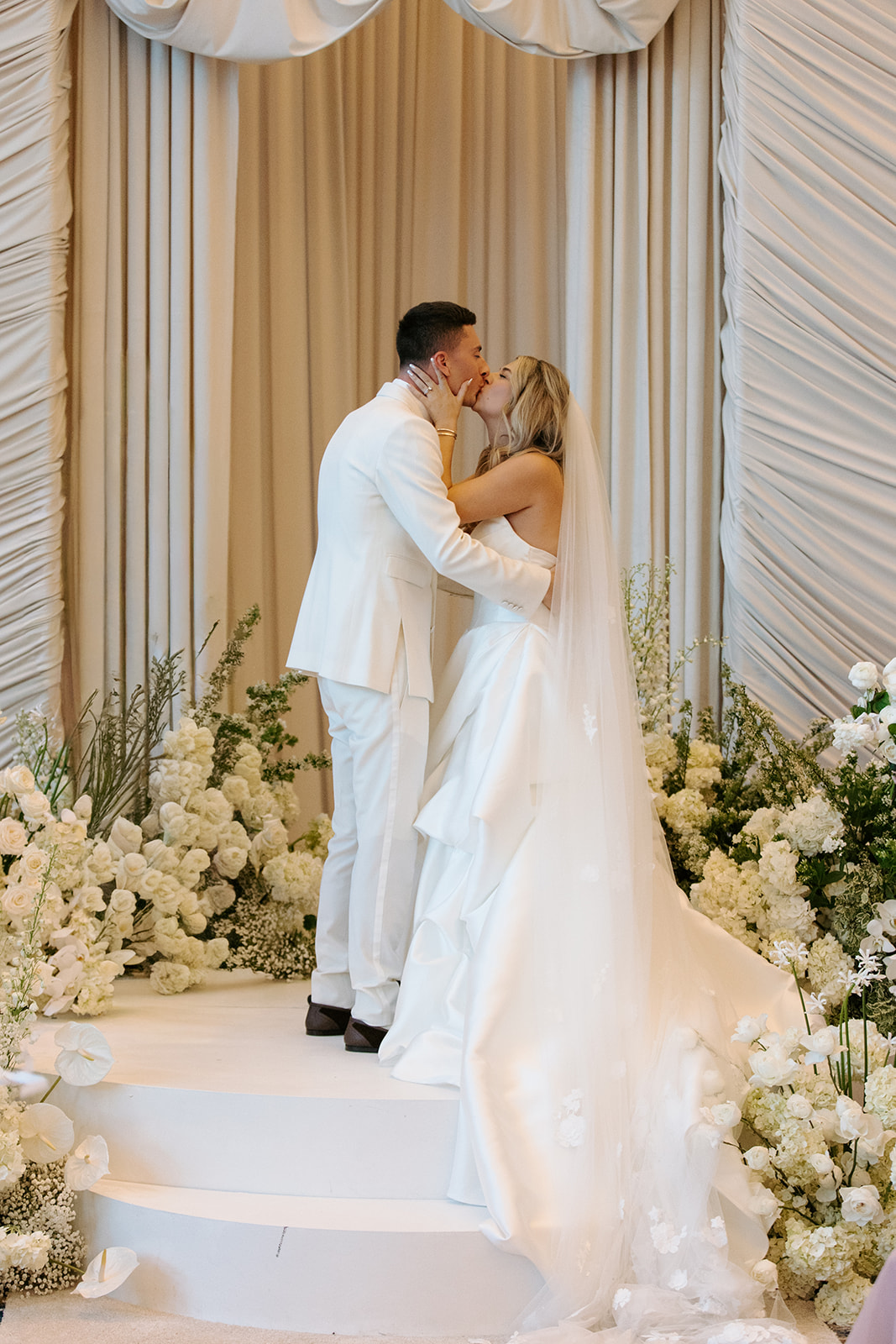 A couple in wedding attire kissing on a decorated stage with white flowers and draped curtains.
