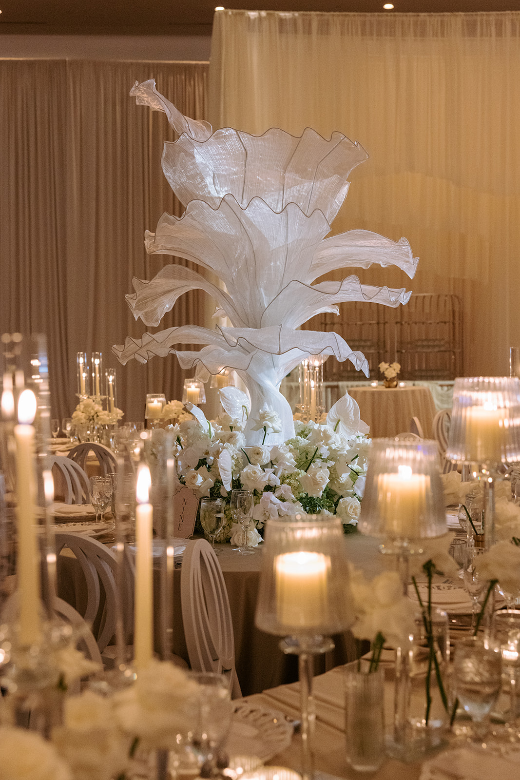Elegant table centerpiece with white flowers and large white decorative sculpture, surrounded by lit candles and tableware, in a decorated event space.