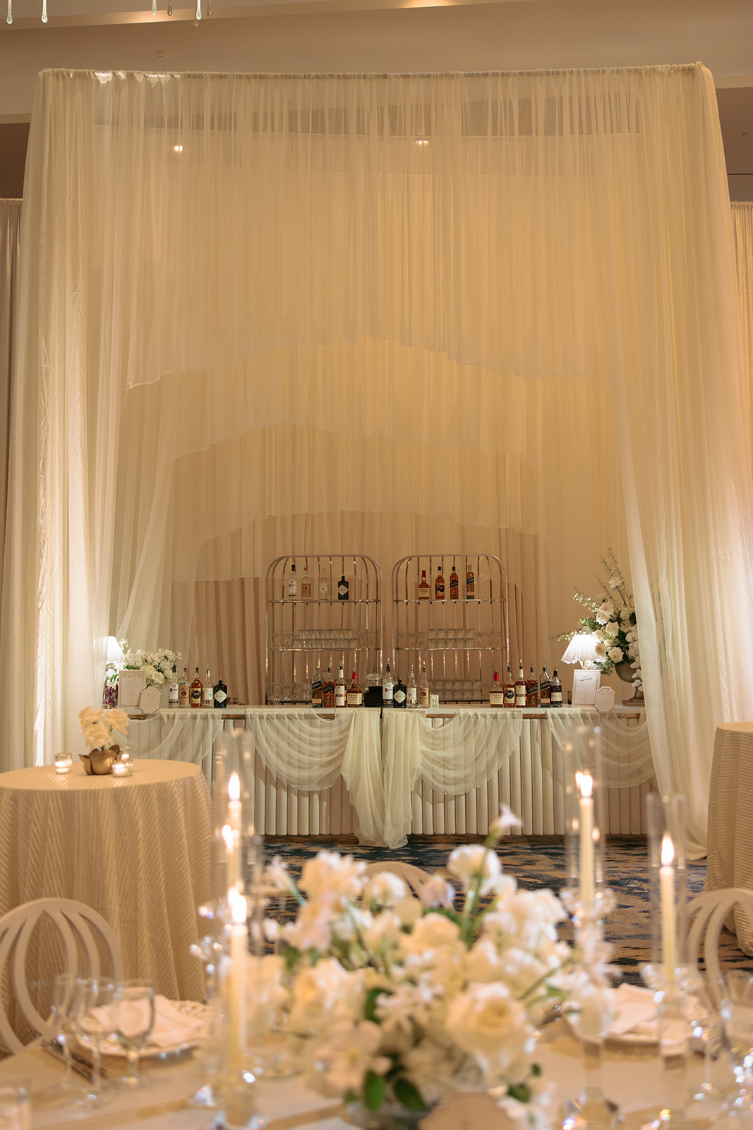 Elegant event setup with draped fabric backdrop, floral arrangements, and table settings in warm lighting.