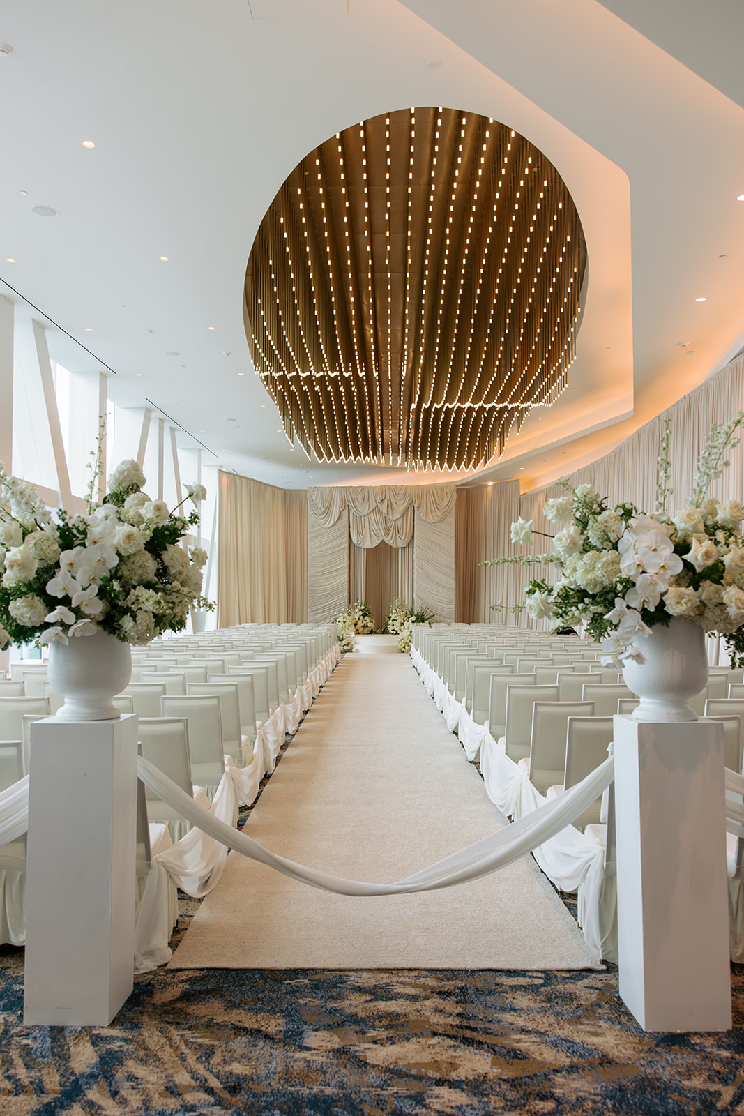 Elegant event space with floral arrangements, white chairs, and a large gold circular ceiling feature.