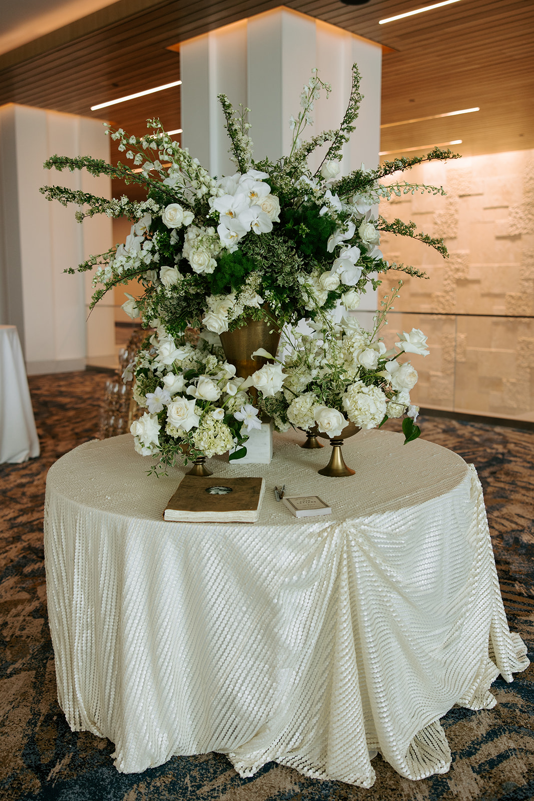 Large floral arrangement with white flowers and greenery on a round table with a cream-colored tablecloth, indoors.