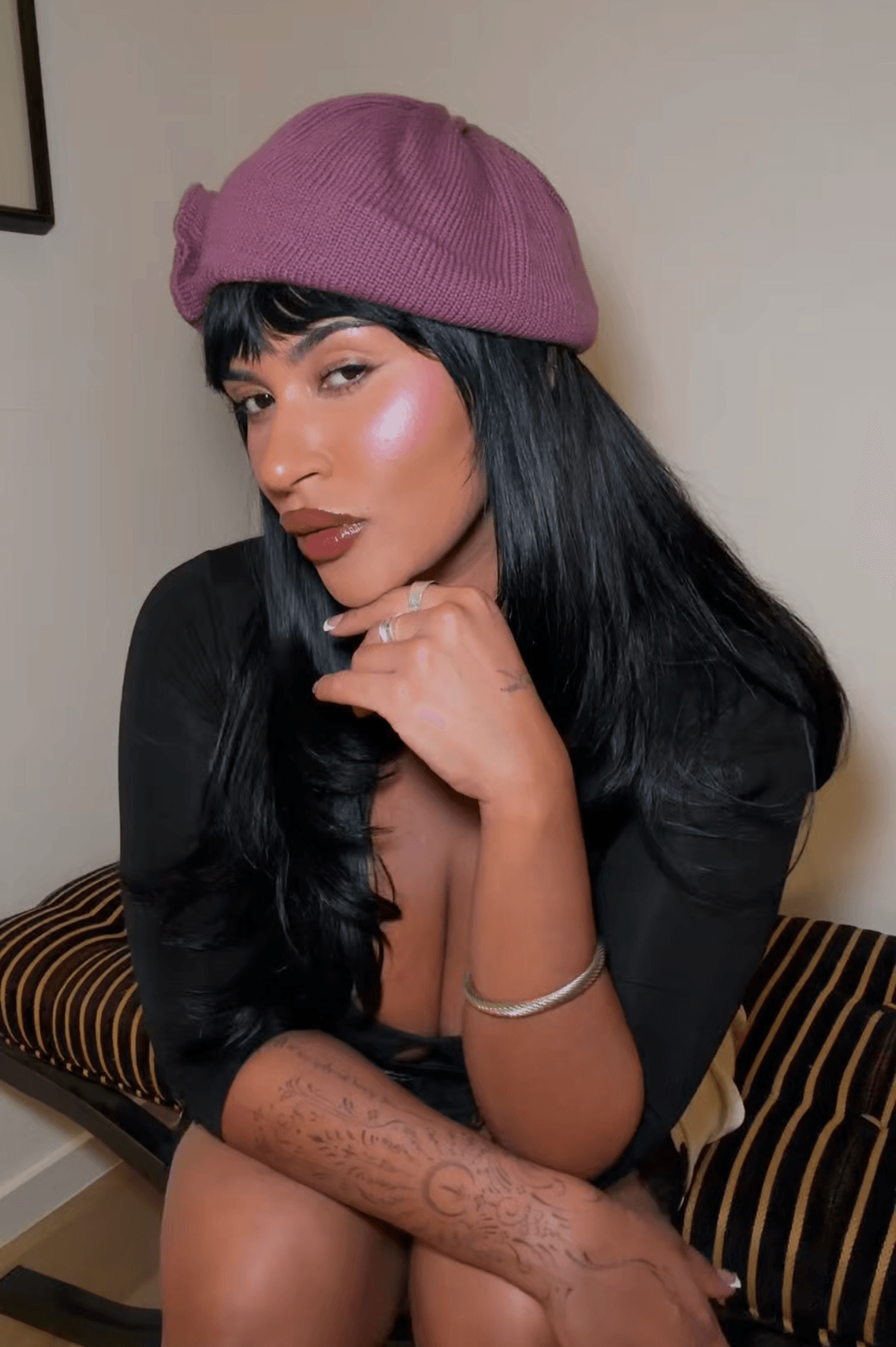 Woman with black hair wearing a pink beret and black top, sitting on a striped cushion, resting her chin on her hand.