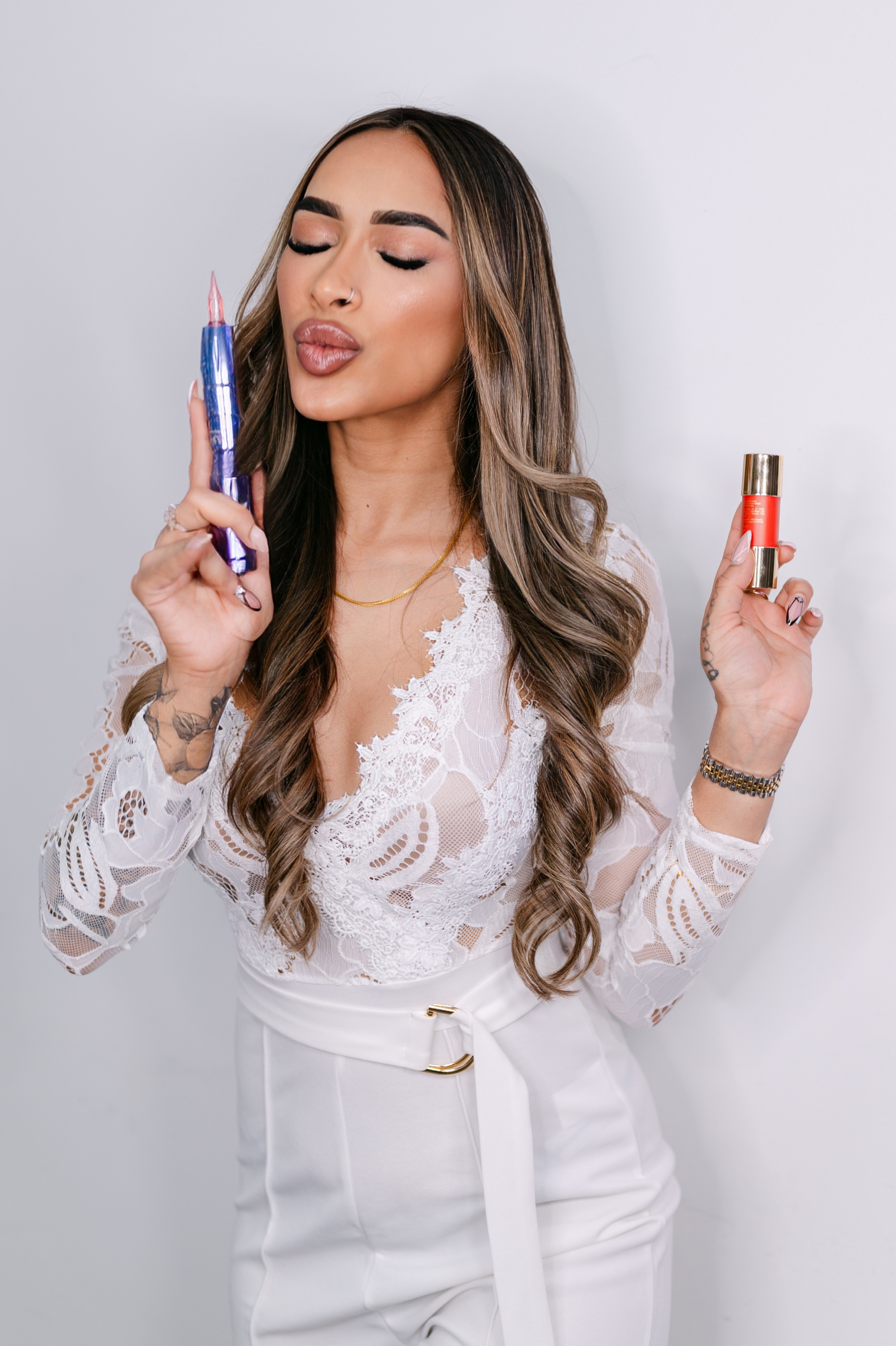 Woman with long wavy hair holding a pen and lipstick, eyes closed, lips puckered, wearing a white lace top.